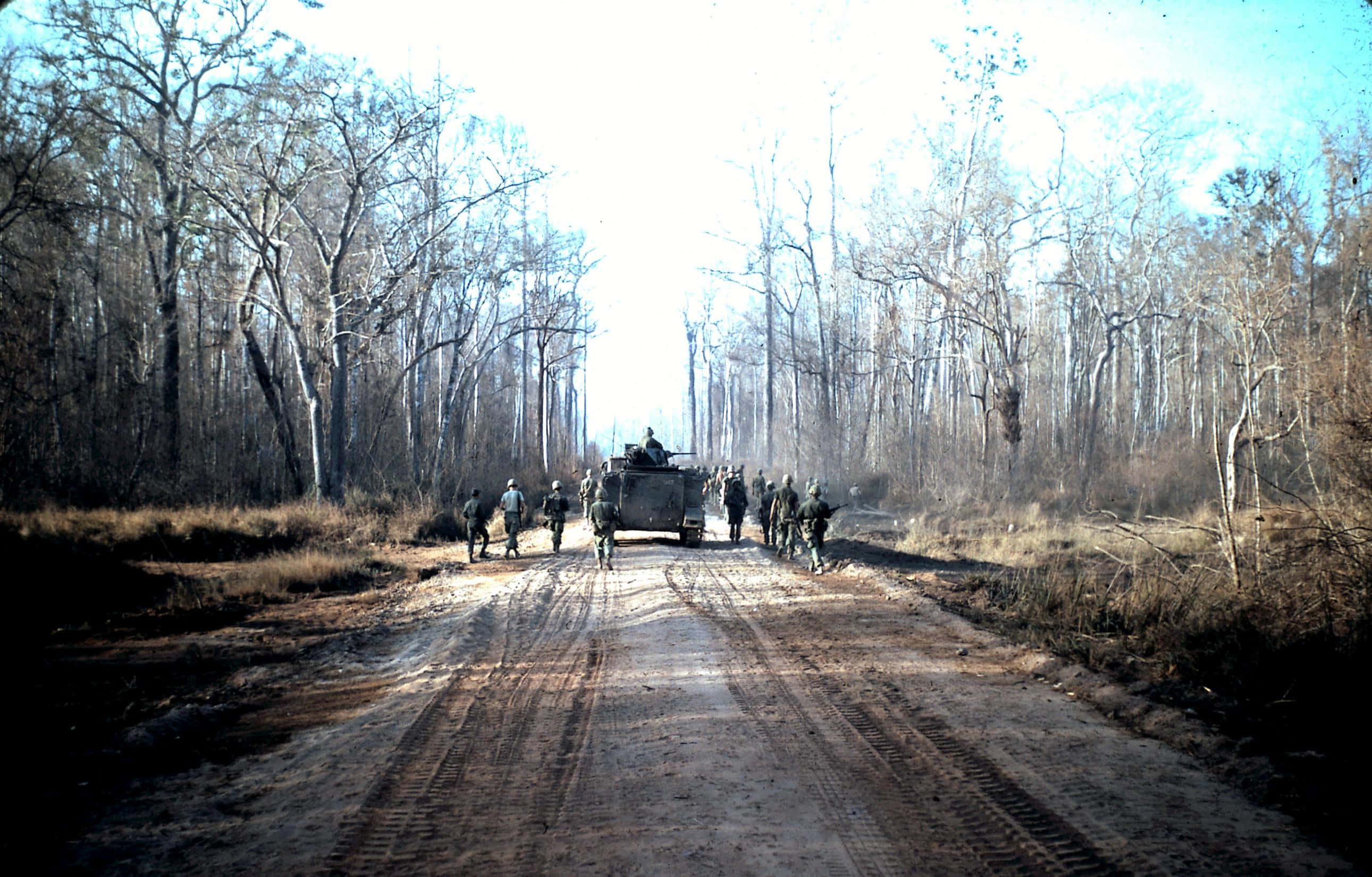 Vietnam War 桌布 壁紙 — 士兵們在明亮的天空下，沿著泥濘的森林道路與軍用車輛並排行進。