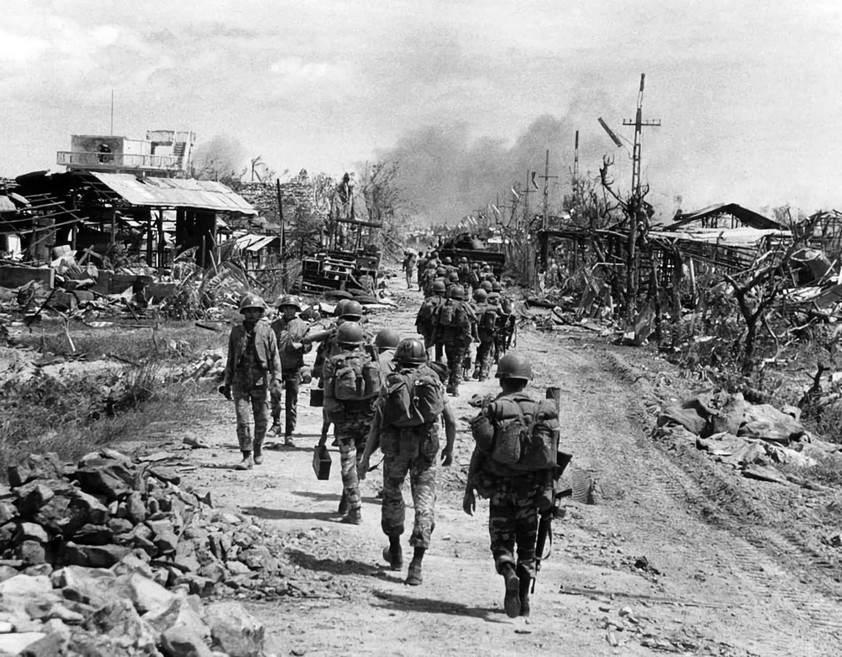 Vietnam War 桌布 壁紙 — 士兵們沿著佈滿瓦礫的道路行進，穿過一個被摧毀的村莊，背景中煙霧升騰。