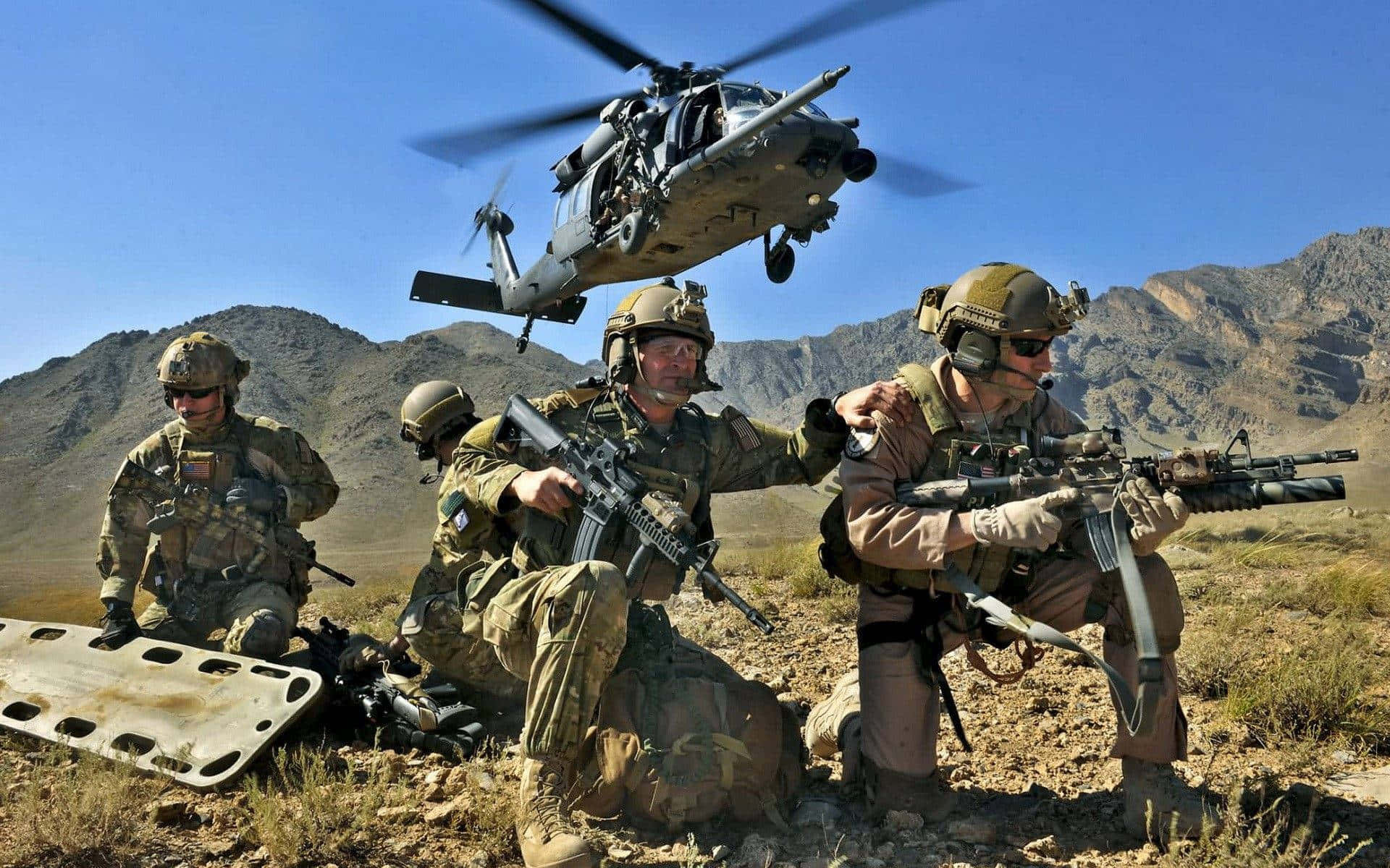 美國陸軍 桌布 壁紙 — 士兵在沙漠地形中，直升機在頭頂盤旋，背景是岩石山脈，天空清澈湛藍。