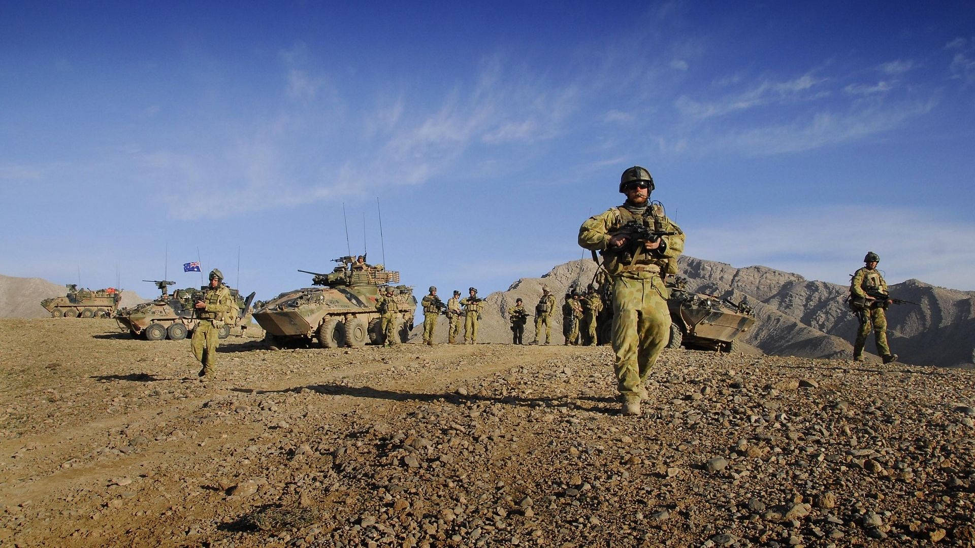 士兵在沙漠山地景觀中與裝甲車輛一同行進。 — 軍隊 壁紙