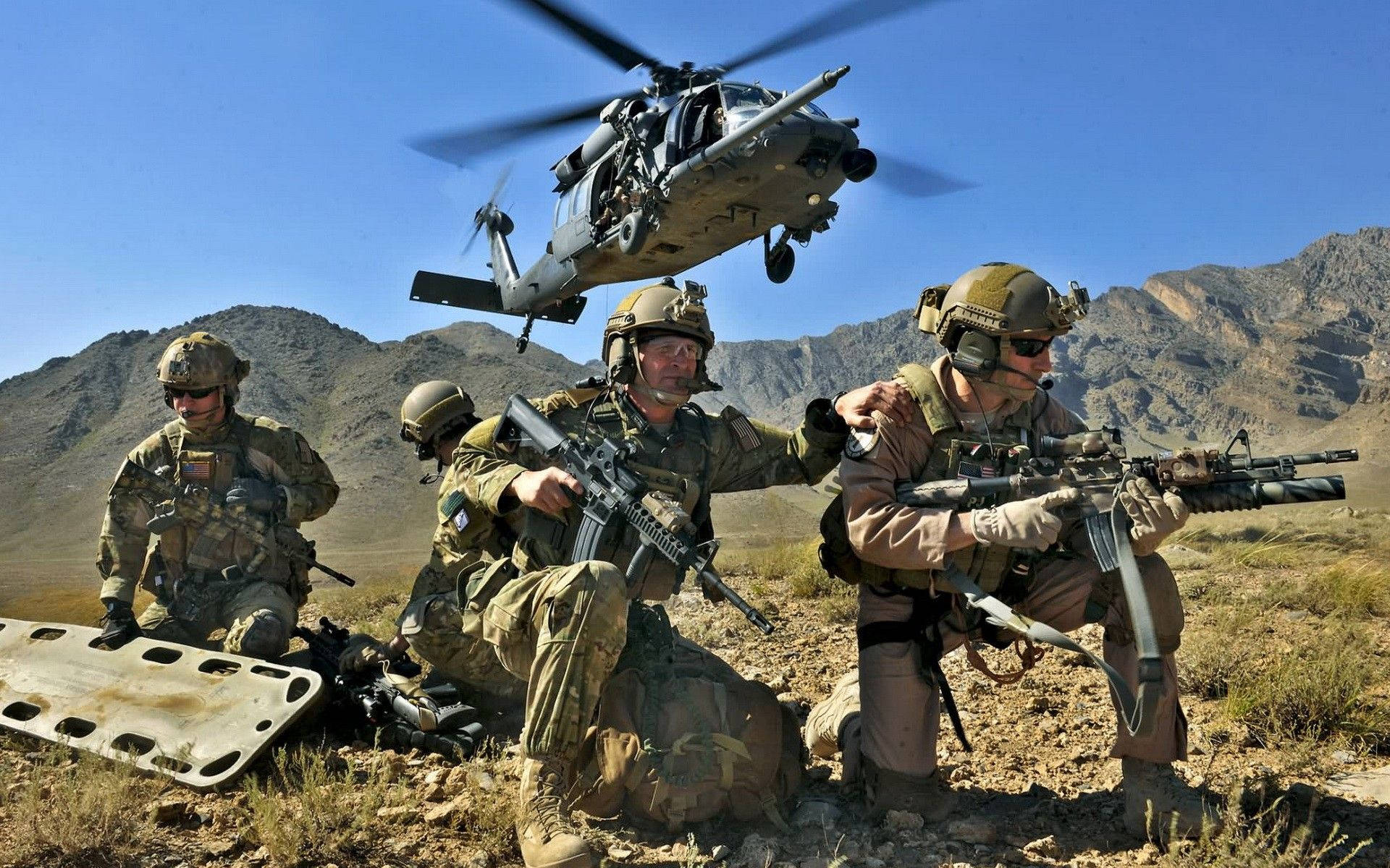 US 桌布 壁紙 — 士兵在沙漠地形中蹲下持步槍，一架軍用直升機在崎嶇山脈上空盤旋。