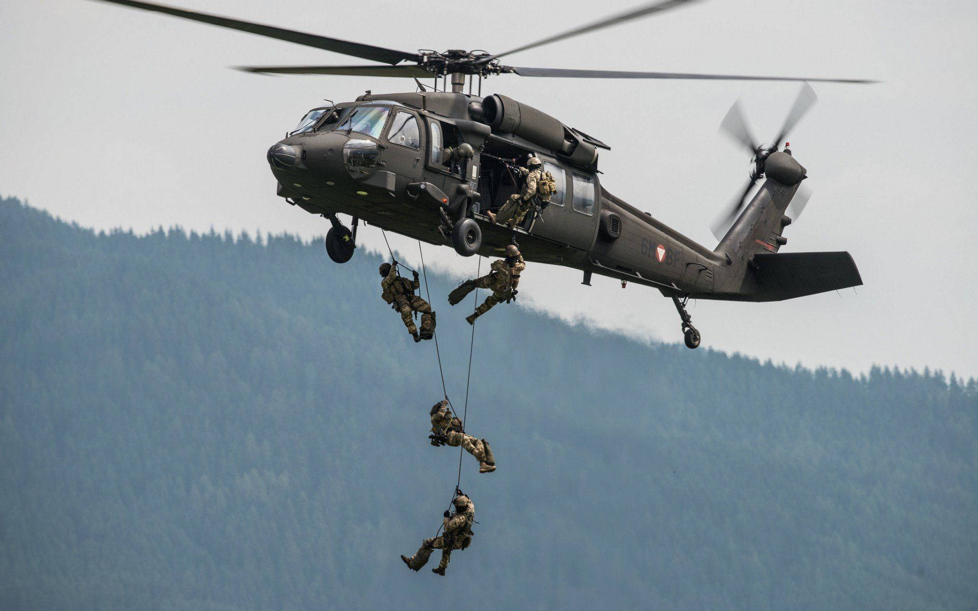 Black Hawk Helicopter 桌布 壁紙 — 士兵從軍用直升機上垂降，背景是陰天下的森林覆蓋山脈。
