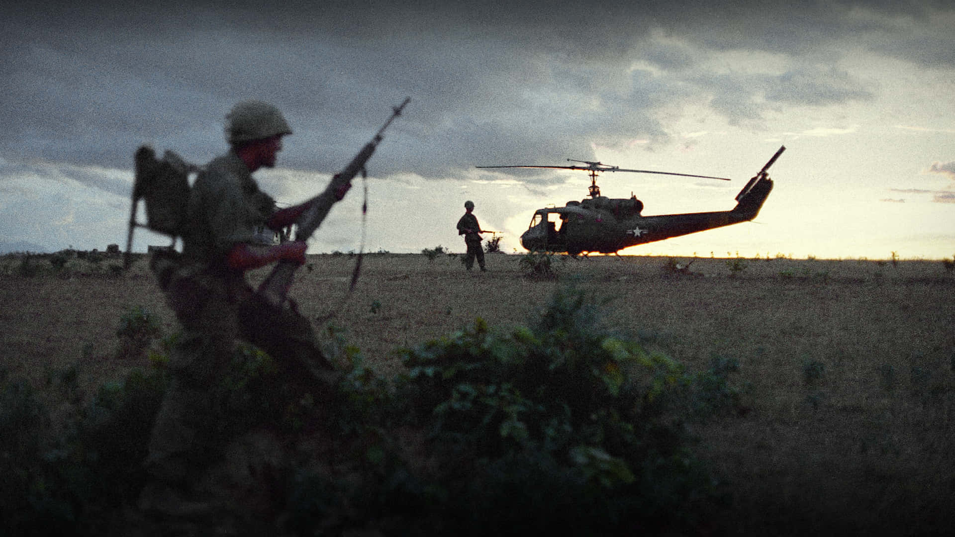 Vietnam War 桌布 壁紙 — 士兵持步槍穿過田野，一架直升機在風暴暮色天空下盤旋。