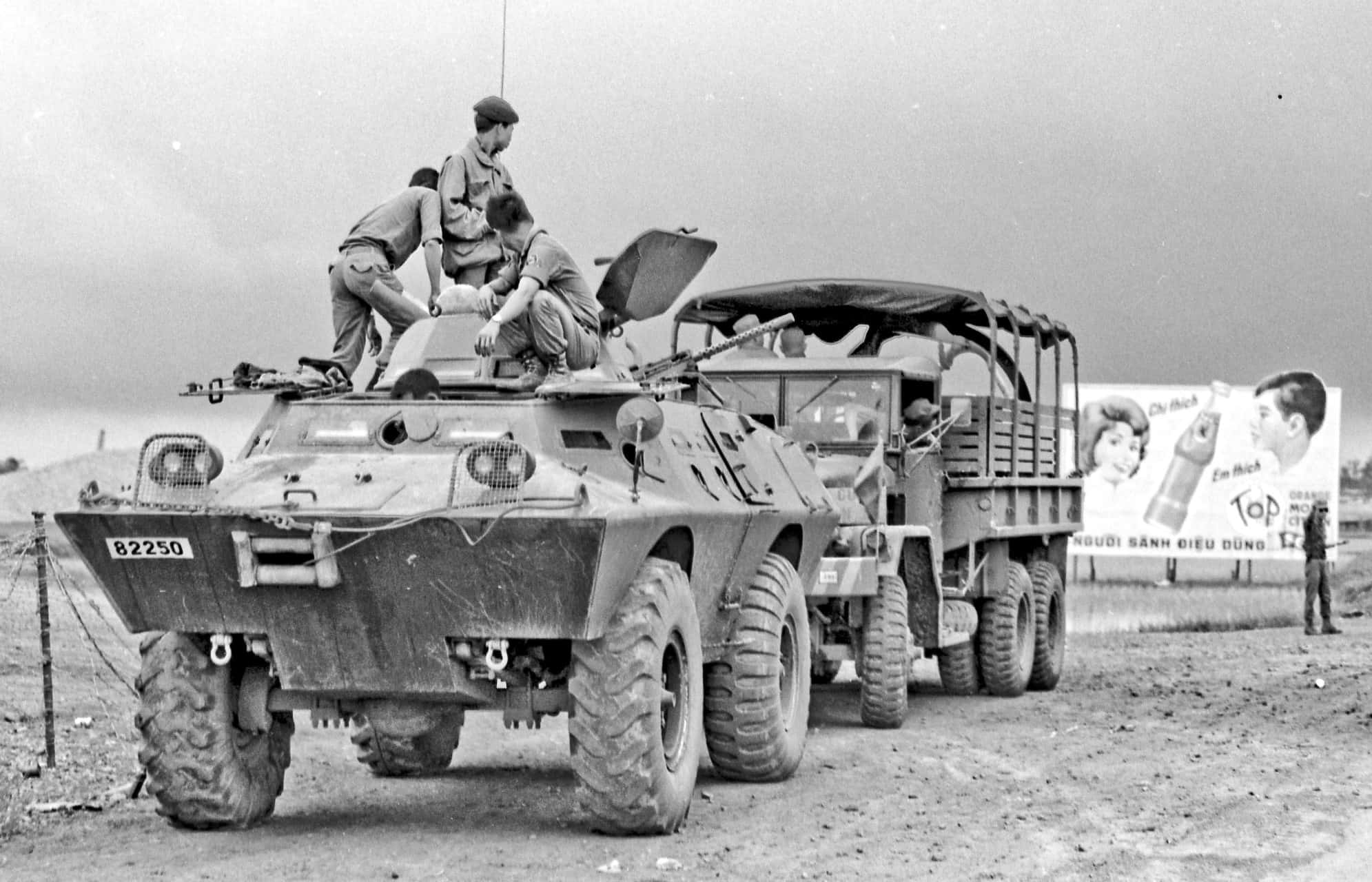 越南 桌布 壁紙 — 士兵爬上停在卡車旁邊的堅固裝甲車，背景有復古廣告看板。