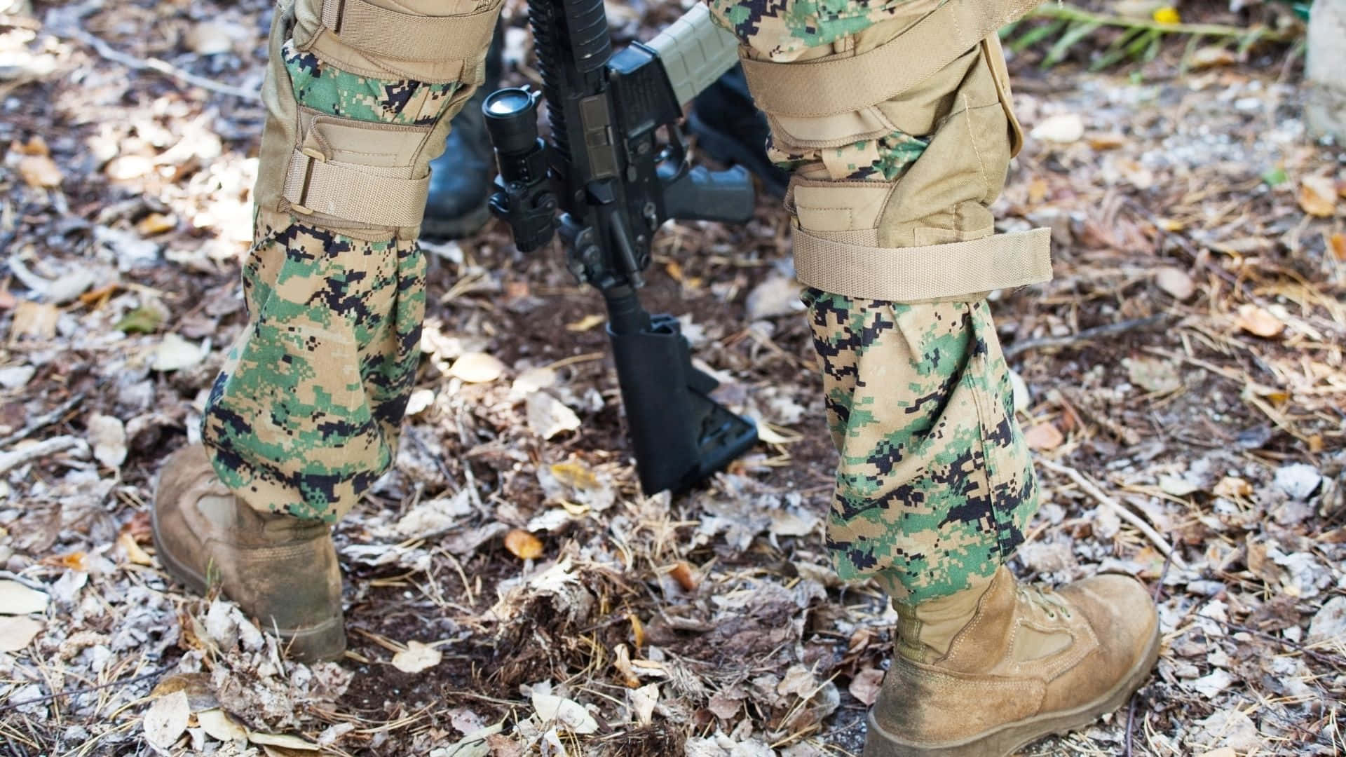 軍事動圖 桌布 壁紙 — 士兵穿著迷彩裝備，在森林地面的落葉中手持步槍的腿部特寫。