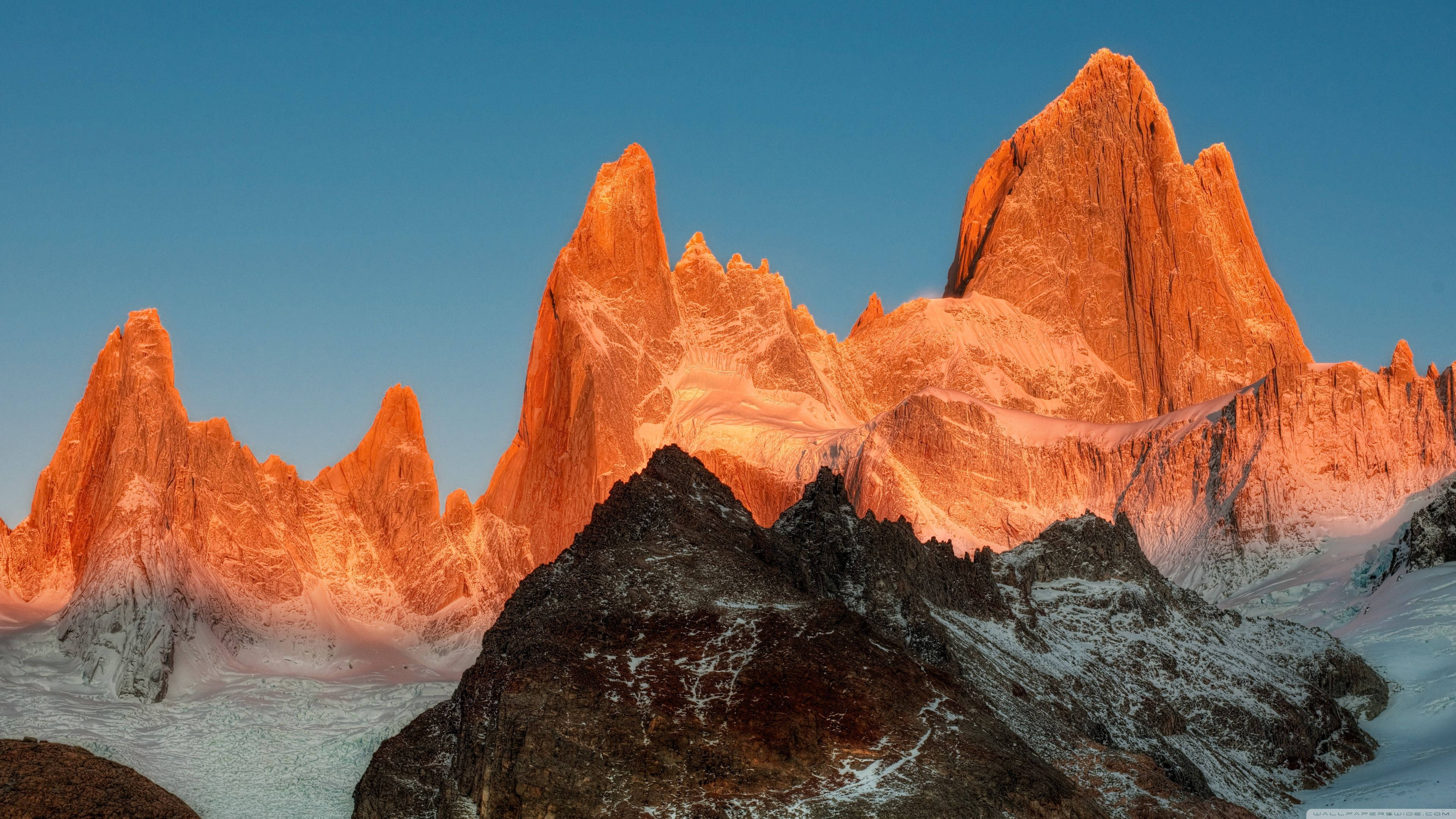 Patagonia 桌布 壁紙 — 壯觀的鋸齒狀山峰在日出時閃耀著火橙色，映襯著深藍色的天空和白雪覆蓋的前景。