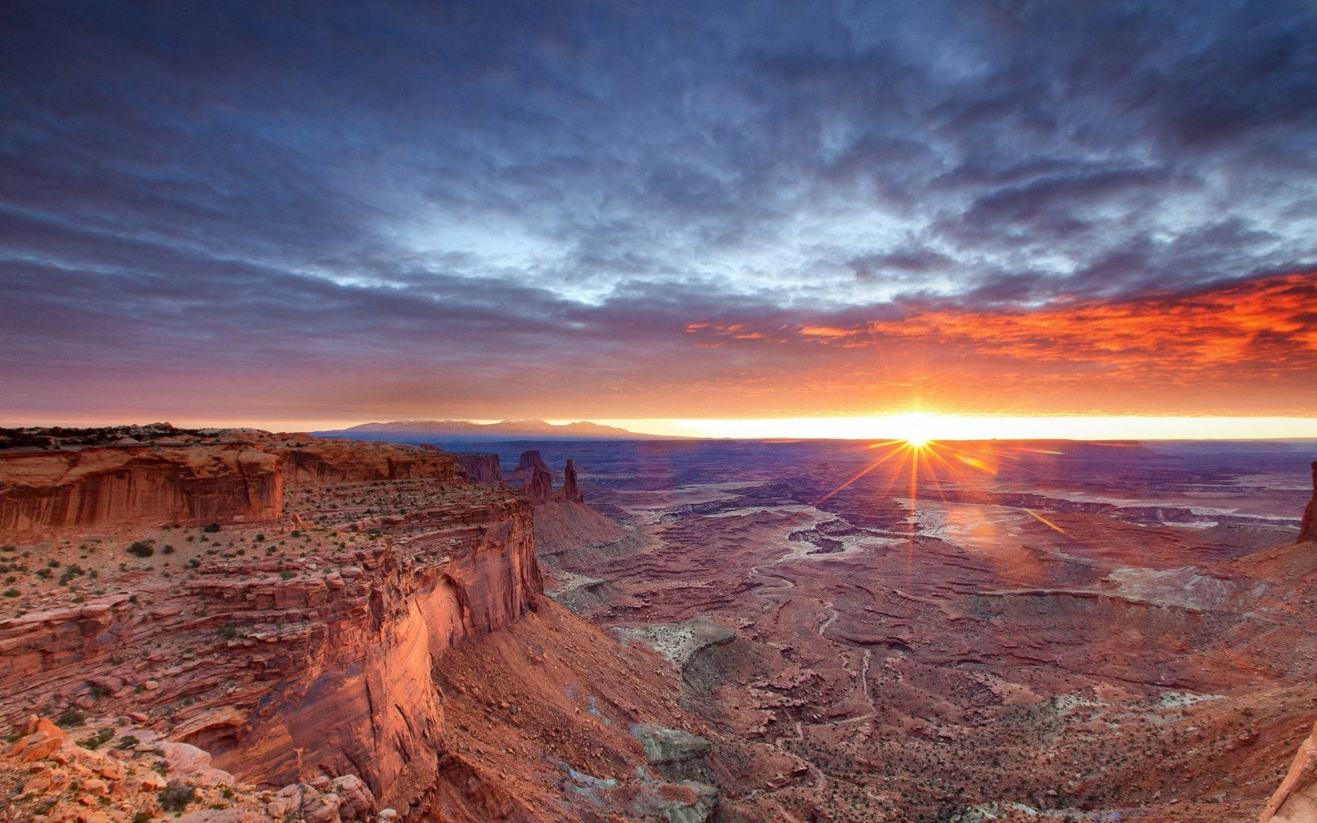 Canyonlands National Park 桌布 壁紙 — 壯麗的日出映照在廣闊峽谷上，層層疊疊的紅色岩層與戲劇性的紫橙色雲彩交織。
