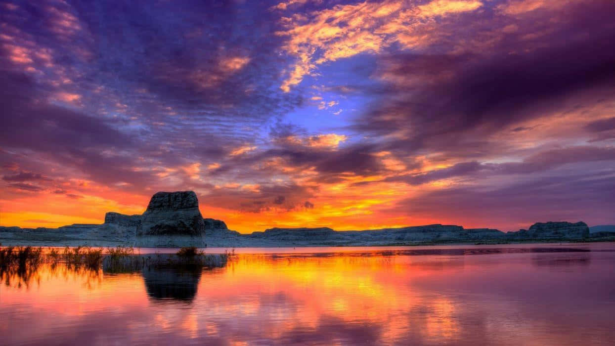 Sunset Landscape 桌布 壁紙 — 壯麗的日落將天空染成火橙色和深紫色，映照在平靜的湖面上，反射出多彩的雲彩和遠處的岩層。