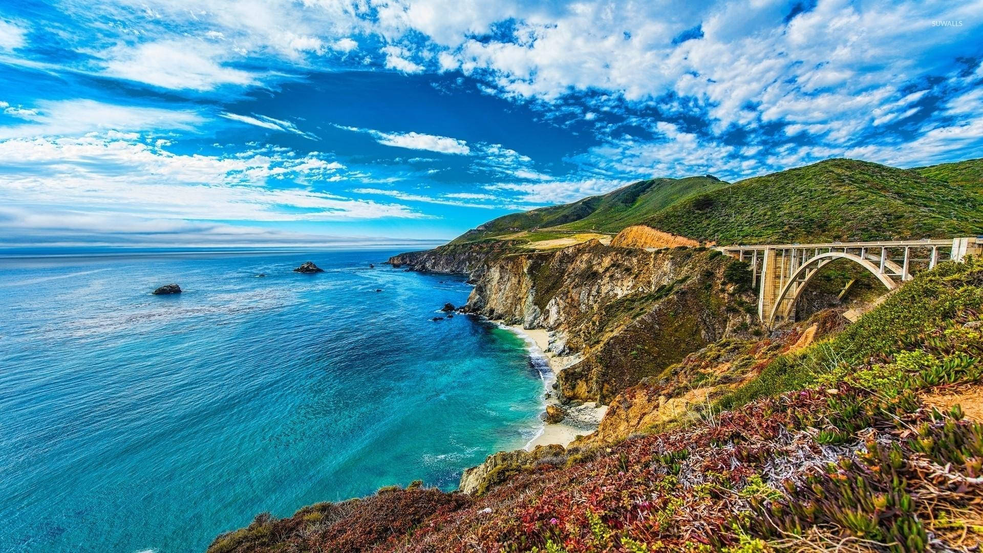 Cloud Aesthetic 桌布 壁紙 — 壯麗的海岸景觀，標誌性的比克斯比橋橫跨崎嶇的懸崖，下方是碧綠的海水，上方是湛藍的天空和蓬鬆的白雲。