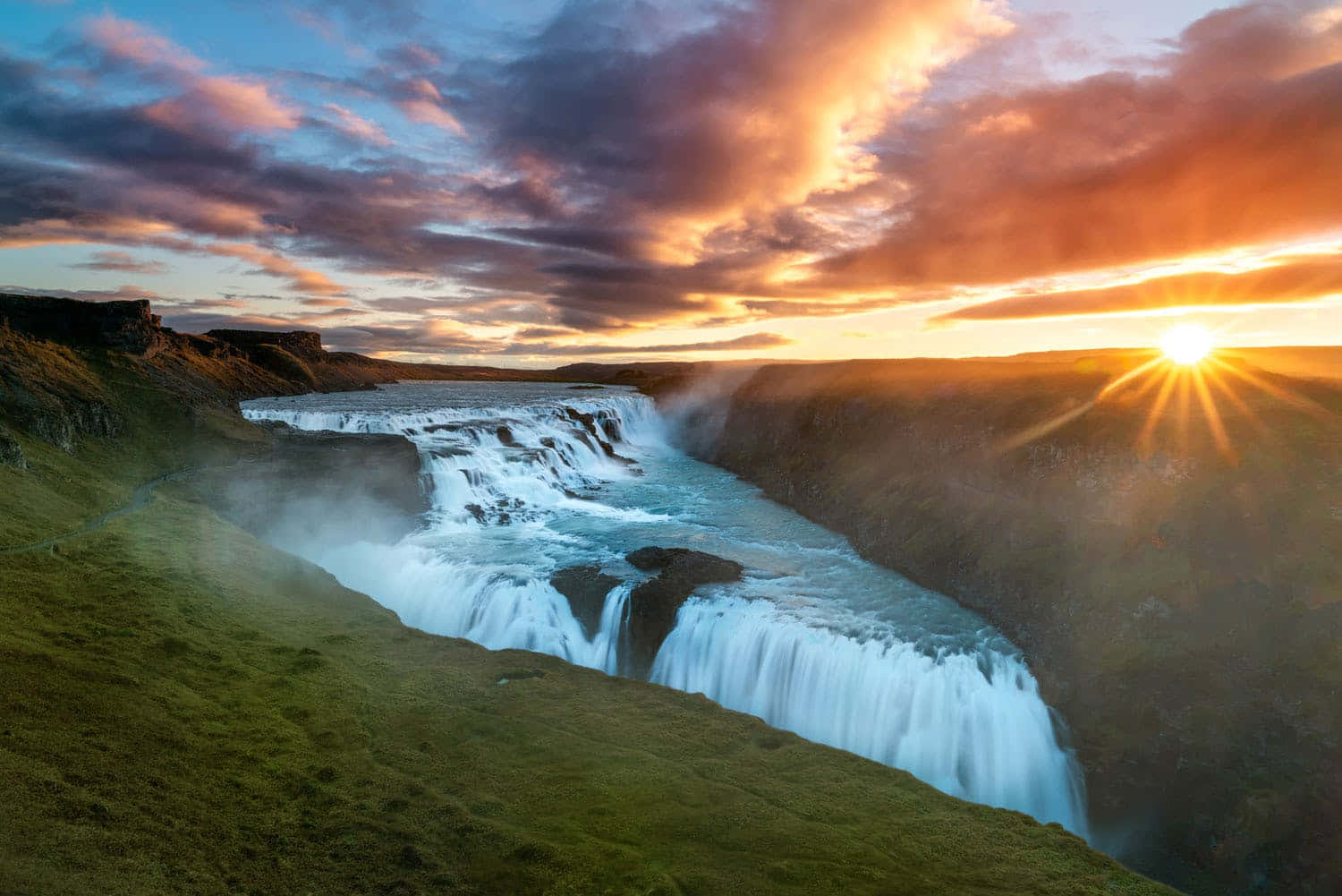 Iceland 桌布 壁紙 — 壯麗的瀑布在日落時分穿過峽谷傾瀉而下，上方有鮮豔的橙色和紫色雲彩。