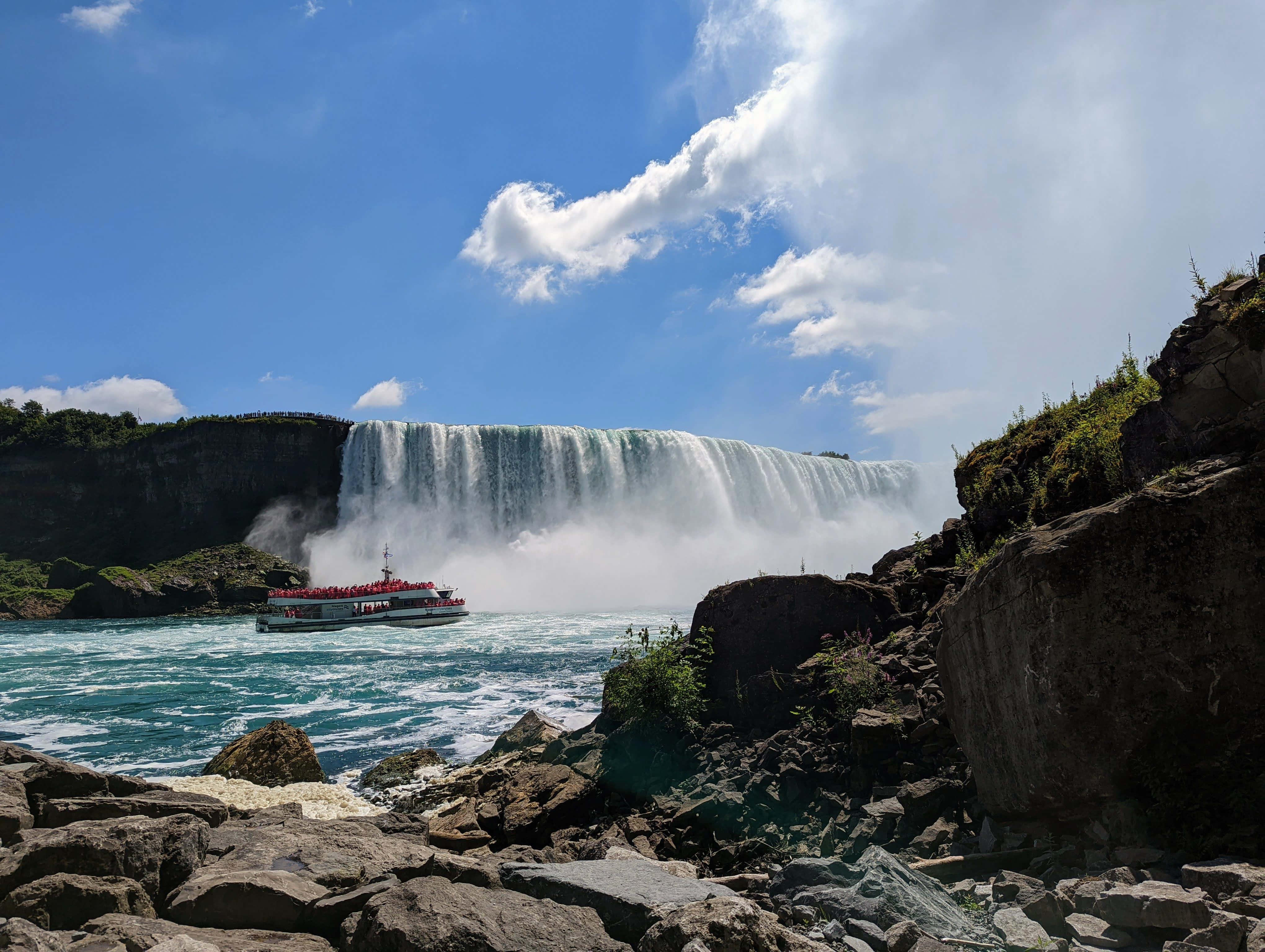 Niagara Falls 桌布 壁紙 — 壯麗的瀑布在明亮的藍天下傾瀉而下，觀光船載著乘客穿過下方霧氣瀰漫的河流。