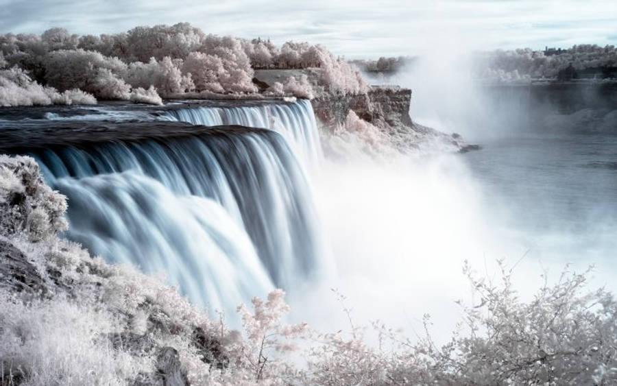 niagara 桌布 壁紙 — 壯麗的瀑布從懸崖上強力傾瀉而下，霧氣升騰，周圍的樹木以超現實的粉紅色和白色色調發光。