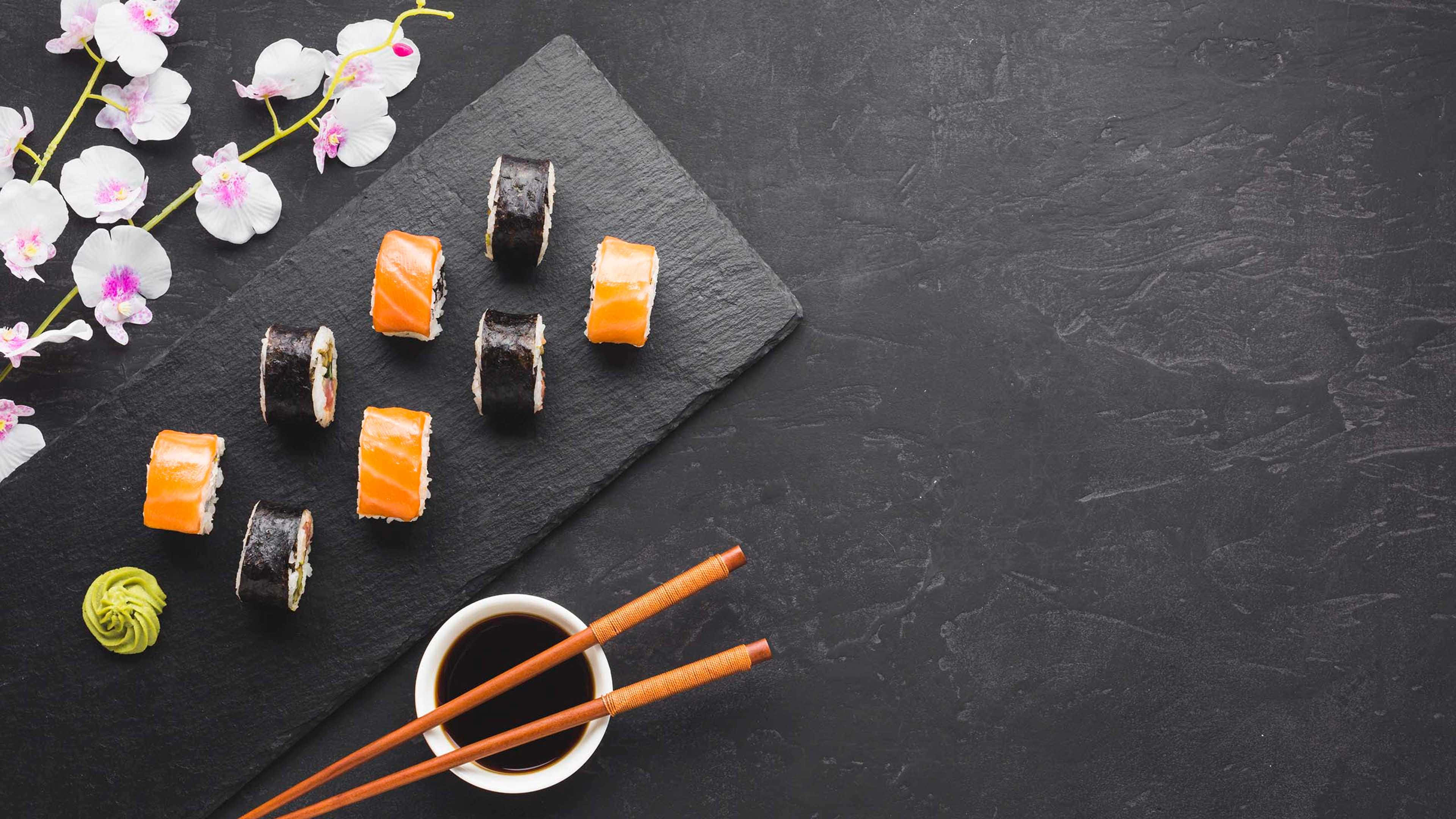 Sushi 桌布 壁紙 — 壽司卷和芥末放在深色石板上的擺盤，搭配蘭花和筷子，背景為有紋理的黑色背景。