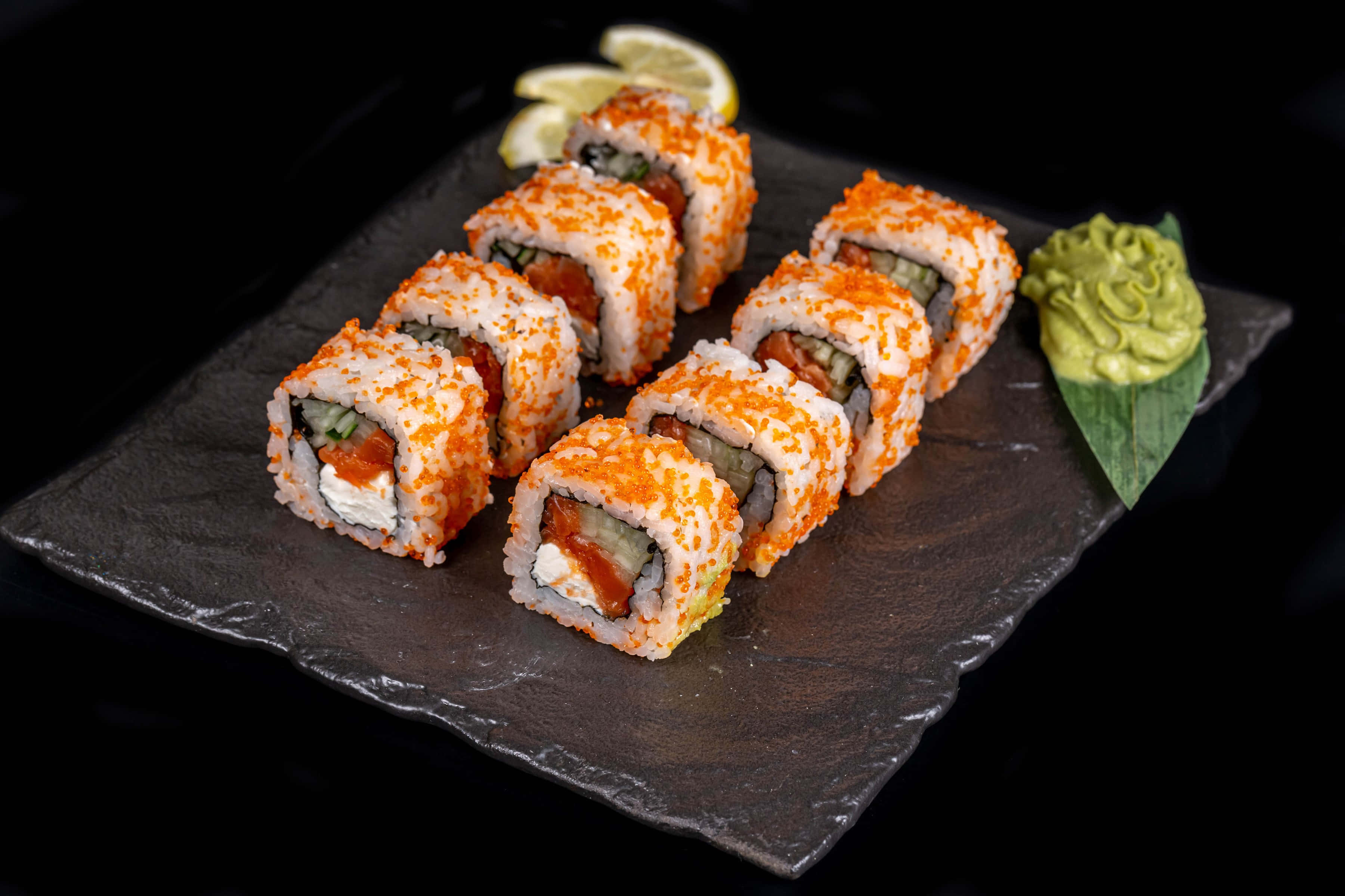 Sushi 桌布 壁紙 — 壽司卷配橙色飛魚籽和奶油餡料，排列在深色石板盤上，搭配芥末和檸檬裝飾。