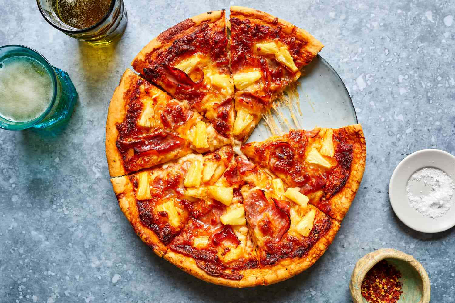 Pizza 桌布 壁紙 — 夏威夷披薩配培根和鳳梨，切片放在盤子上，旁邊有兩瓶啤酒和調味料，置於灰色石面上。