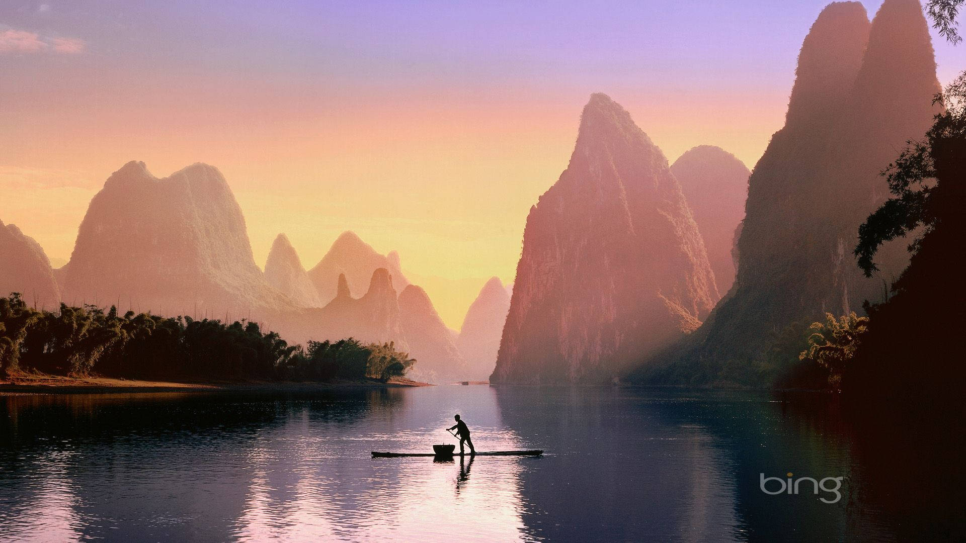 必應 桌布 壁紙 — 夕陽下，一位孤獨的漁夫划著竹筏穿過如鏡的河流，四周是高聳的喀斯特山峰，沐浴在金色的光芒中。