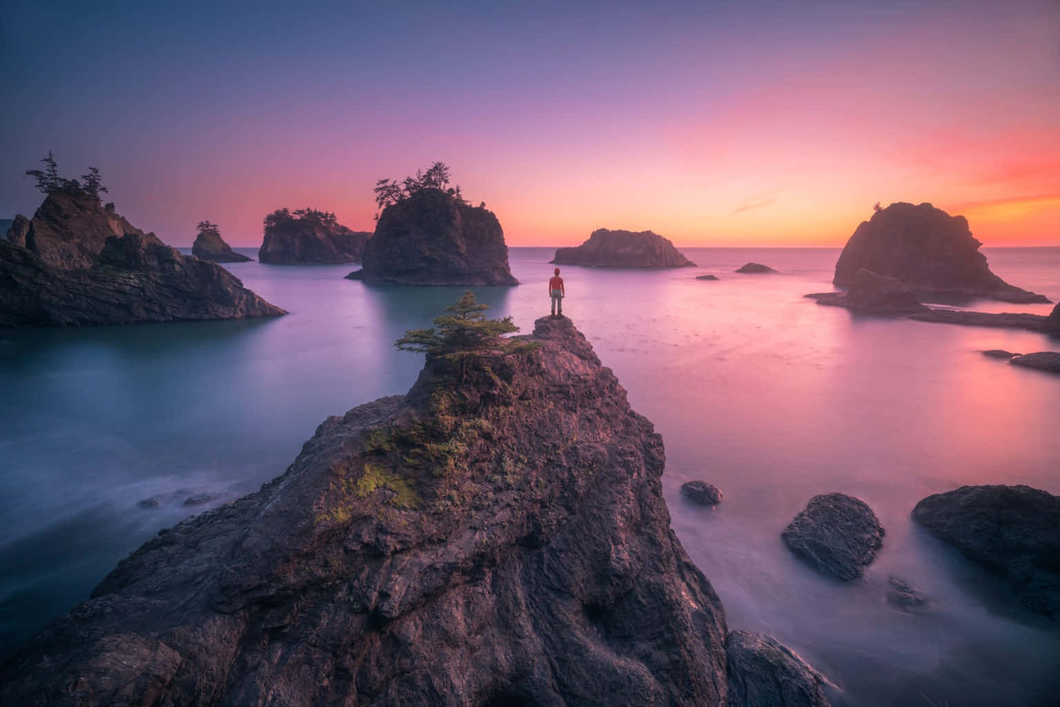 Oregon Coast 桌布 壁紙 — 夕陽下，一個人影獨自站在崎嶇的海岸岩石上，四周是平靜的海水和遠處的小島，天空呈現鮮豔的粉紅色和紫色。