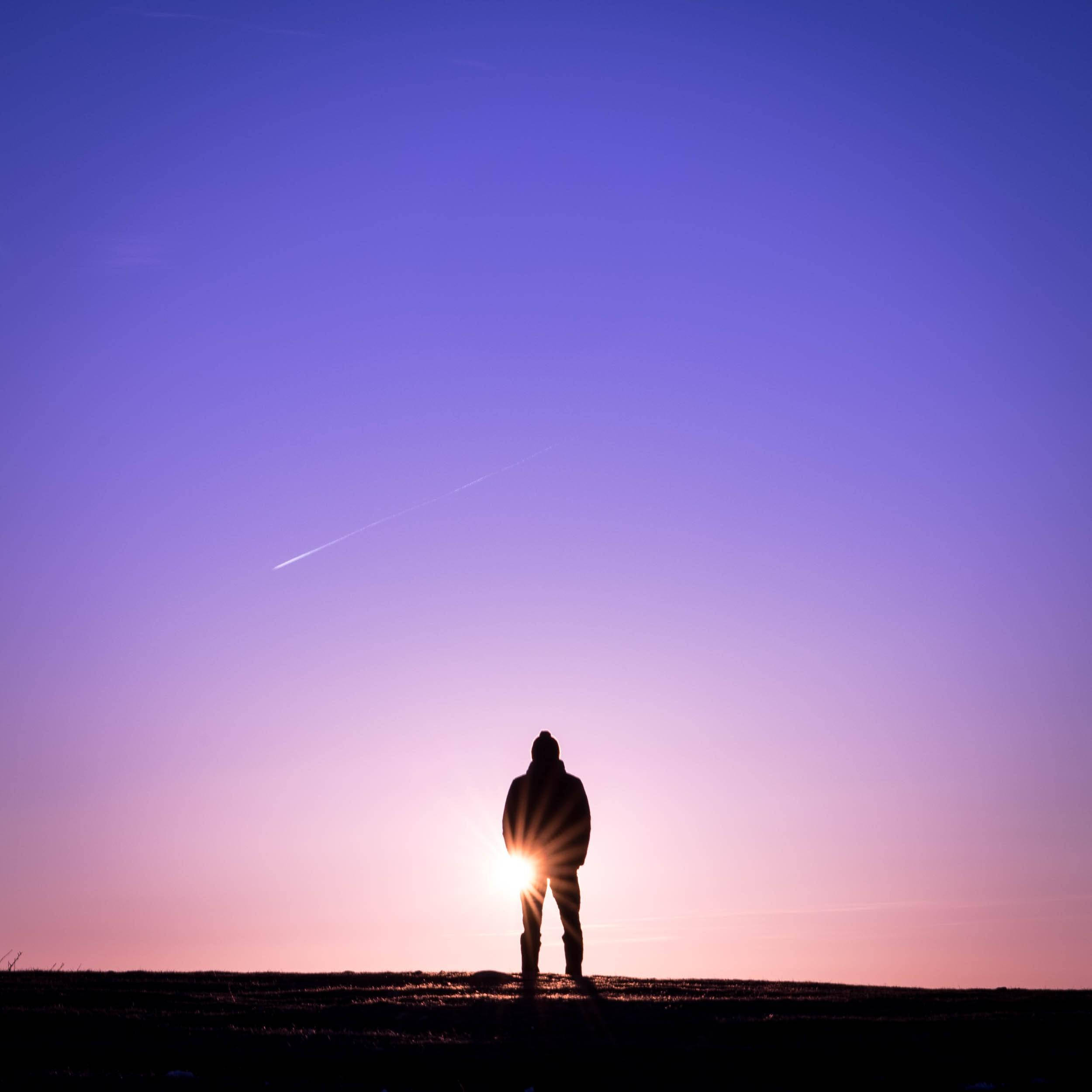 Silhouette 桌布 壁紙 — 夕陽下，一個孤獨的身影站在山丘上，背光襯托著漸變紫色和粉紅色的天空，有一道淡淡的飛機尾跡。