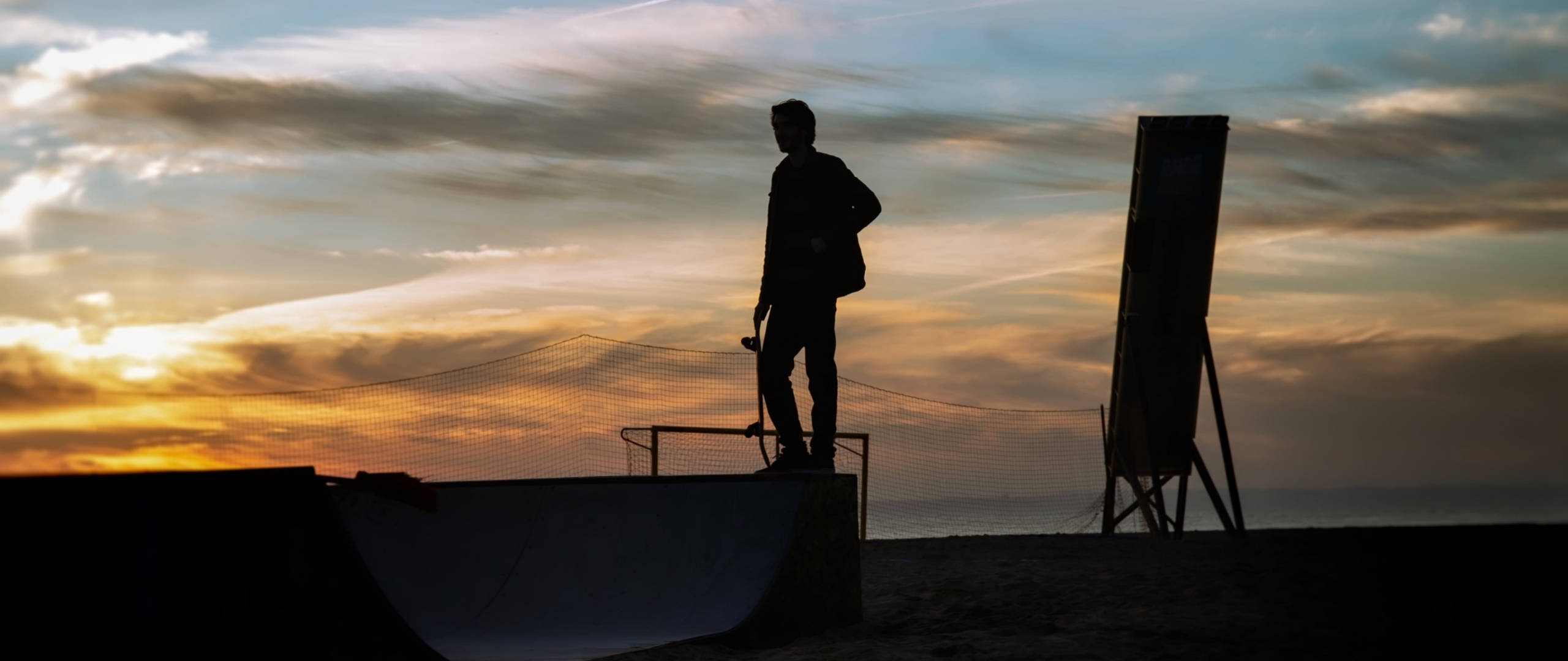 Skater Boy 桌布 壁紙 — 夕陽下，一名滑板手站在斜坡頂端，背後是充滿活力的橙色和藍色天空的剪影。
