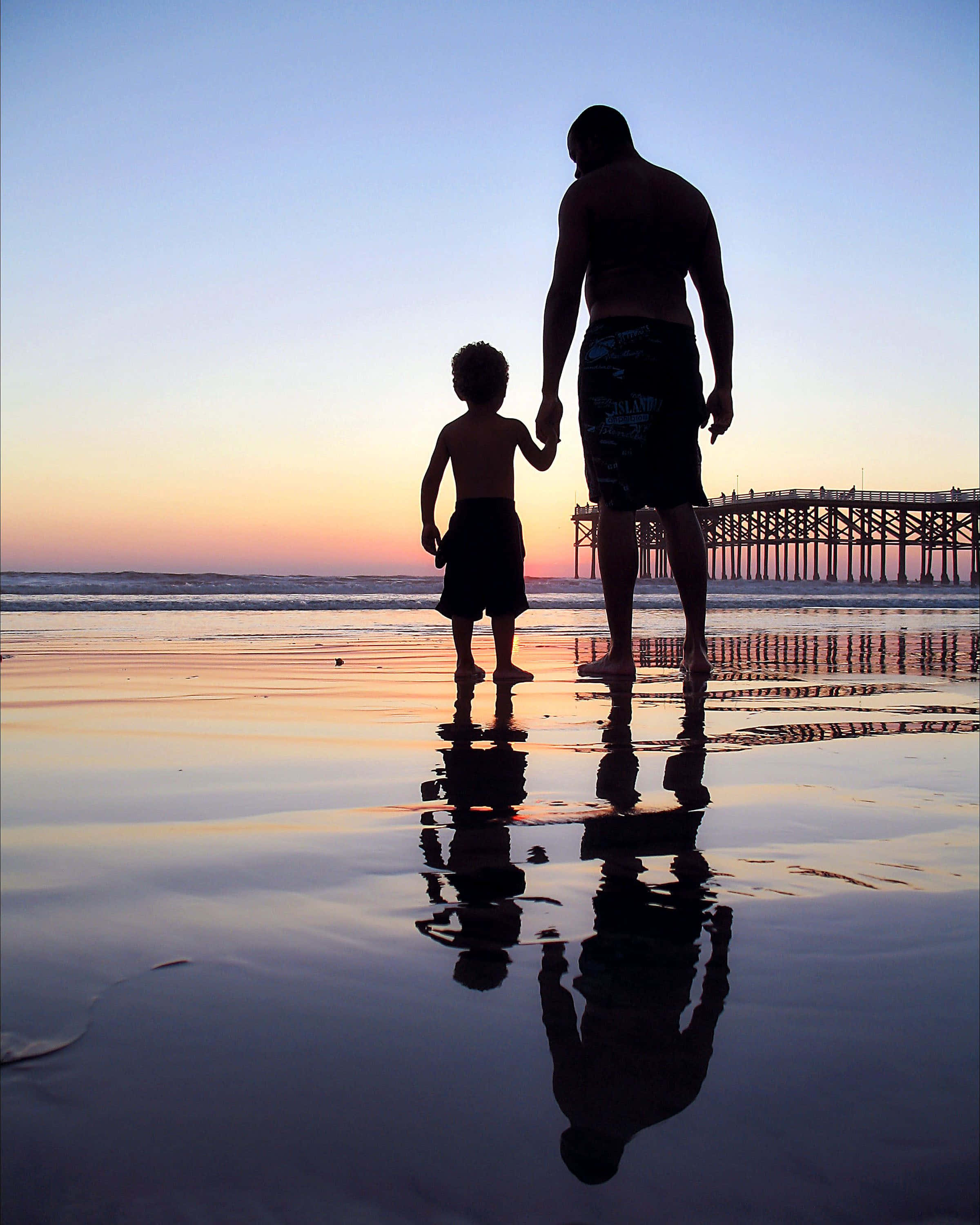Father And Son 桌布 壁紙 — 夕陽下，濕漉漉的海灘上，父親和孩子手牽手的剪影，水中倒映著碼頭。