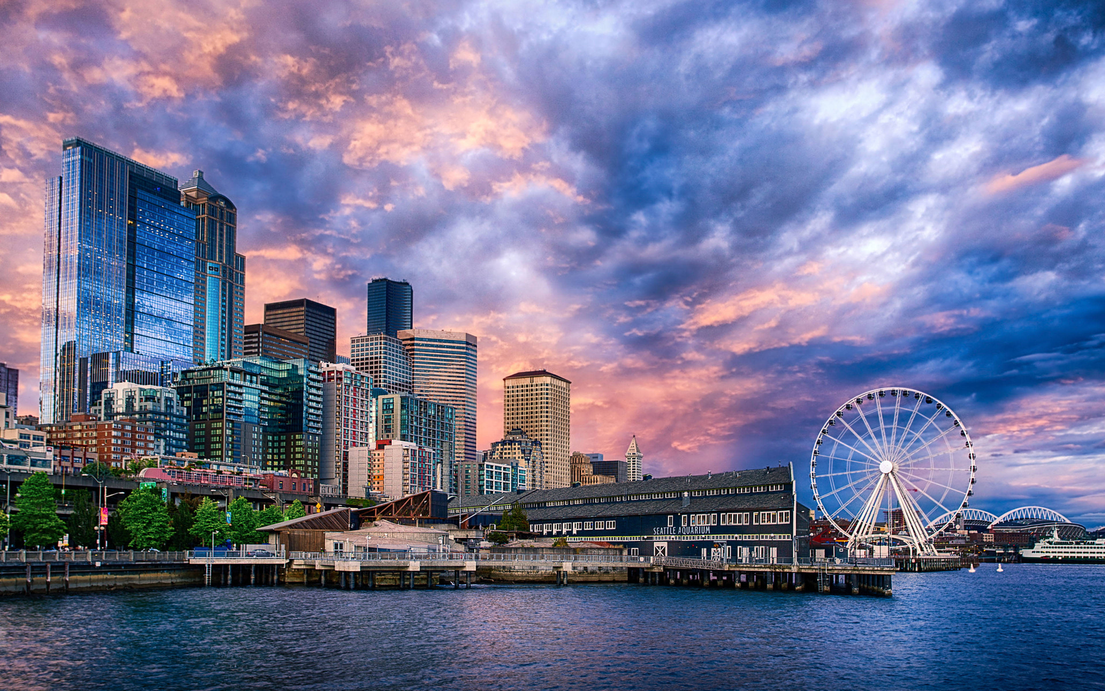 Washington 桌布 壁紙 — 夕陽下充滿活力的城市天際線，海濱有一個巨大的摩天輪，天空佈滿戲劇性的粉紅色和紫色雲彩。