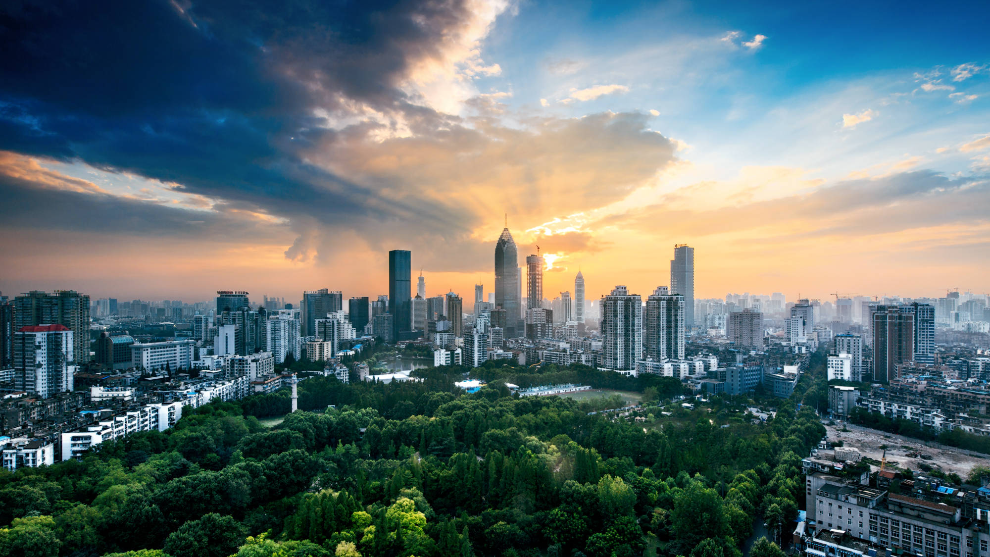 Wuhan 桌布 壁紙 — 夕陽下充滿活力的城市天際線，金色光芒穿透戲劇性的雲層，照耀在鬱鬱蔥蔥的綠色公園之上。