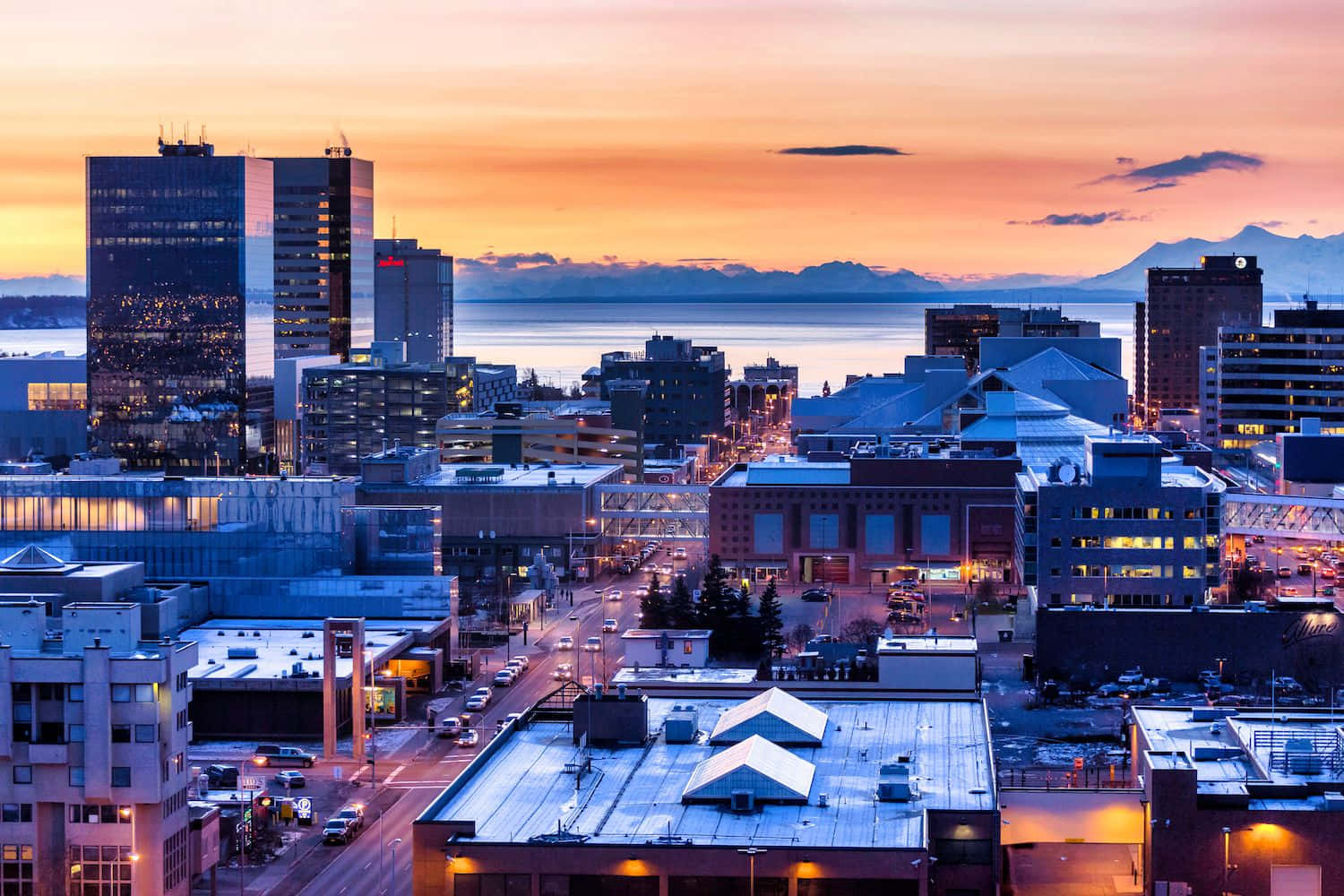 Anchorage Alaska 桌布 壁紙 — 夕陽下充滿活力的城市景觀，發光的摩天大樓和遠處的山脈倒映在平靜的水面上。