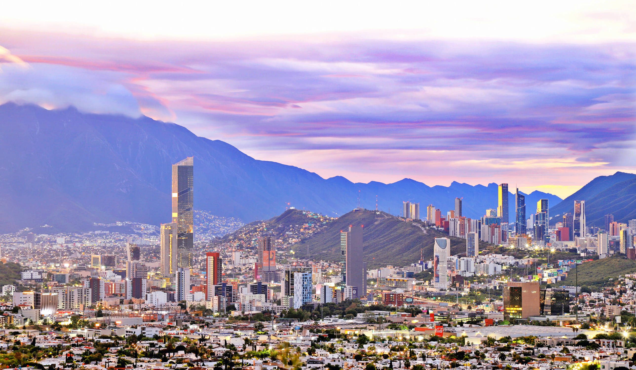 Monterrey 桌布 壁紙 — 夕陽下廣闊的城市景觀，高聳的摩天大樓依偎在紫色調的山脈前，天空呈現充滿活力的粉紅色和薰衣草色。