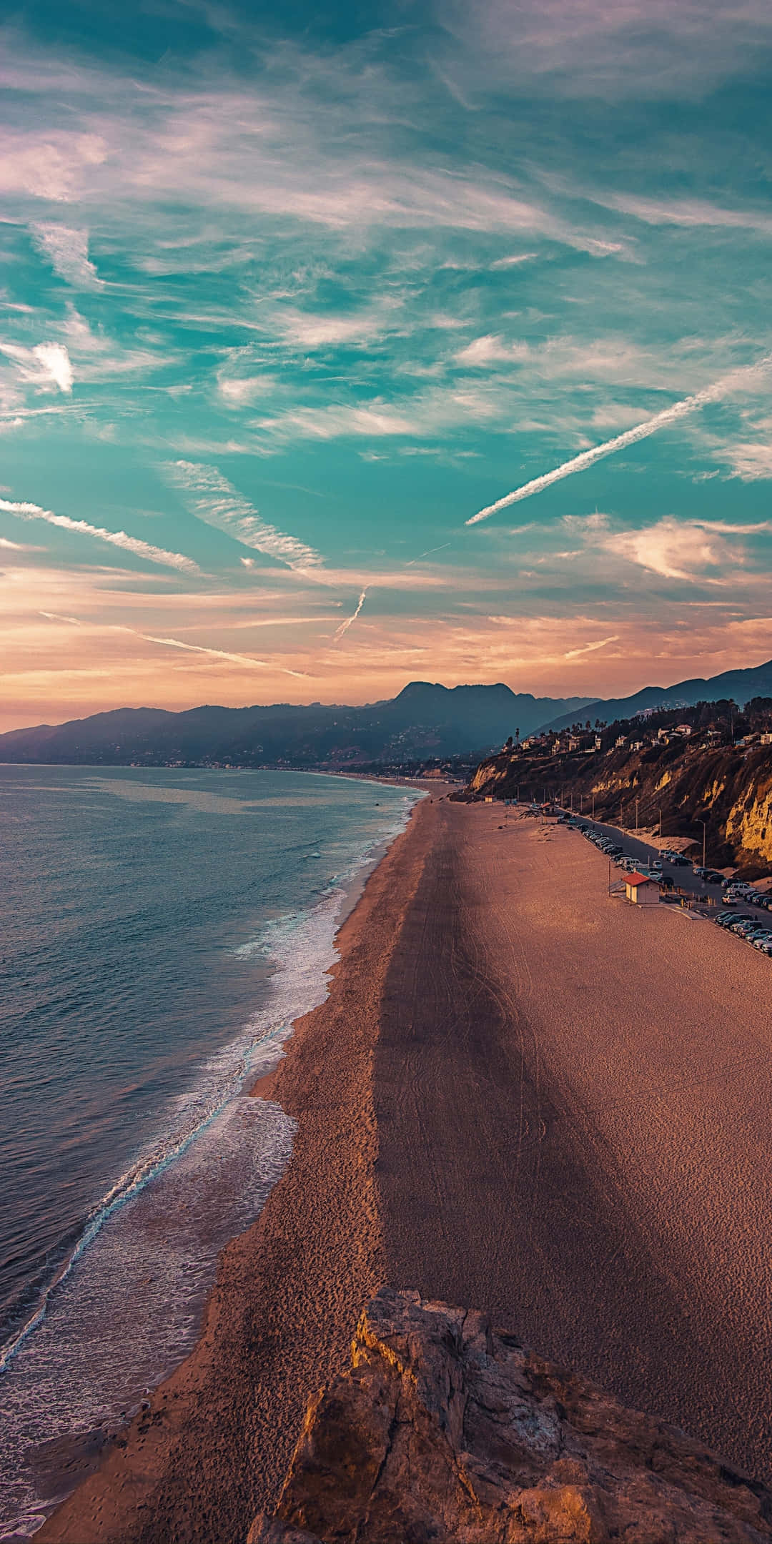 Pixel 3 Beach 桌布 壁紙 — 夕陽下沙灘的鳥瞰圖，碧綠海水、輕柔波浪、遠山，天空帶有條紋雲彩。