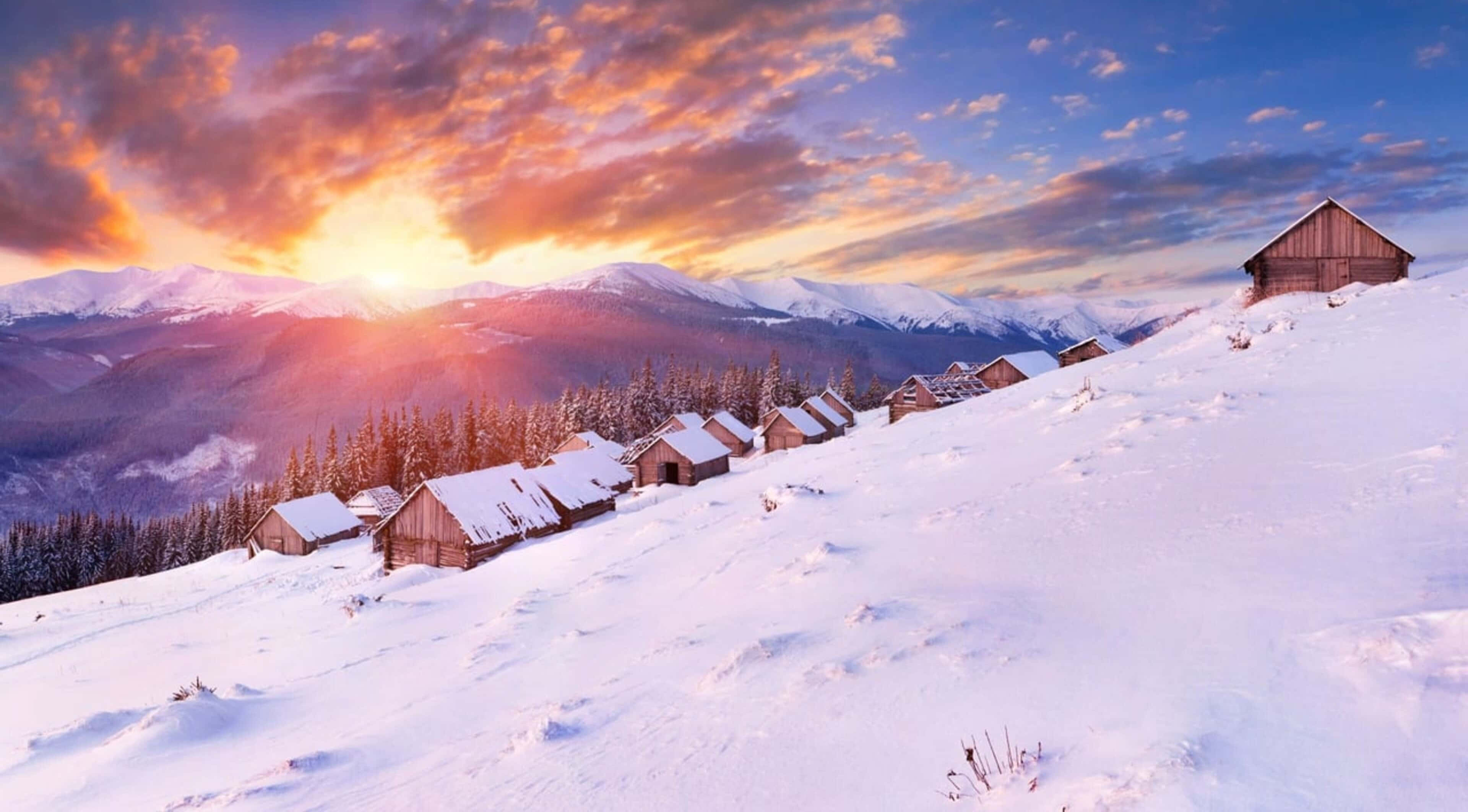 Village 桌布 壁紙 — 夕陽下白雪覆蓋的山村，木屋隱藏在松樹之間，天空呈現火橙色和藍色。