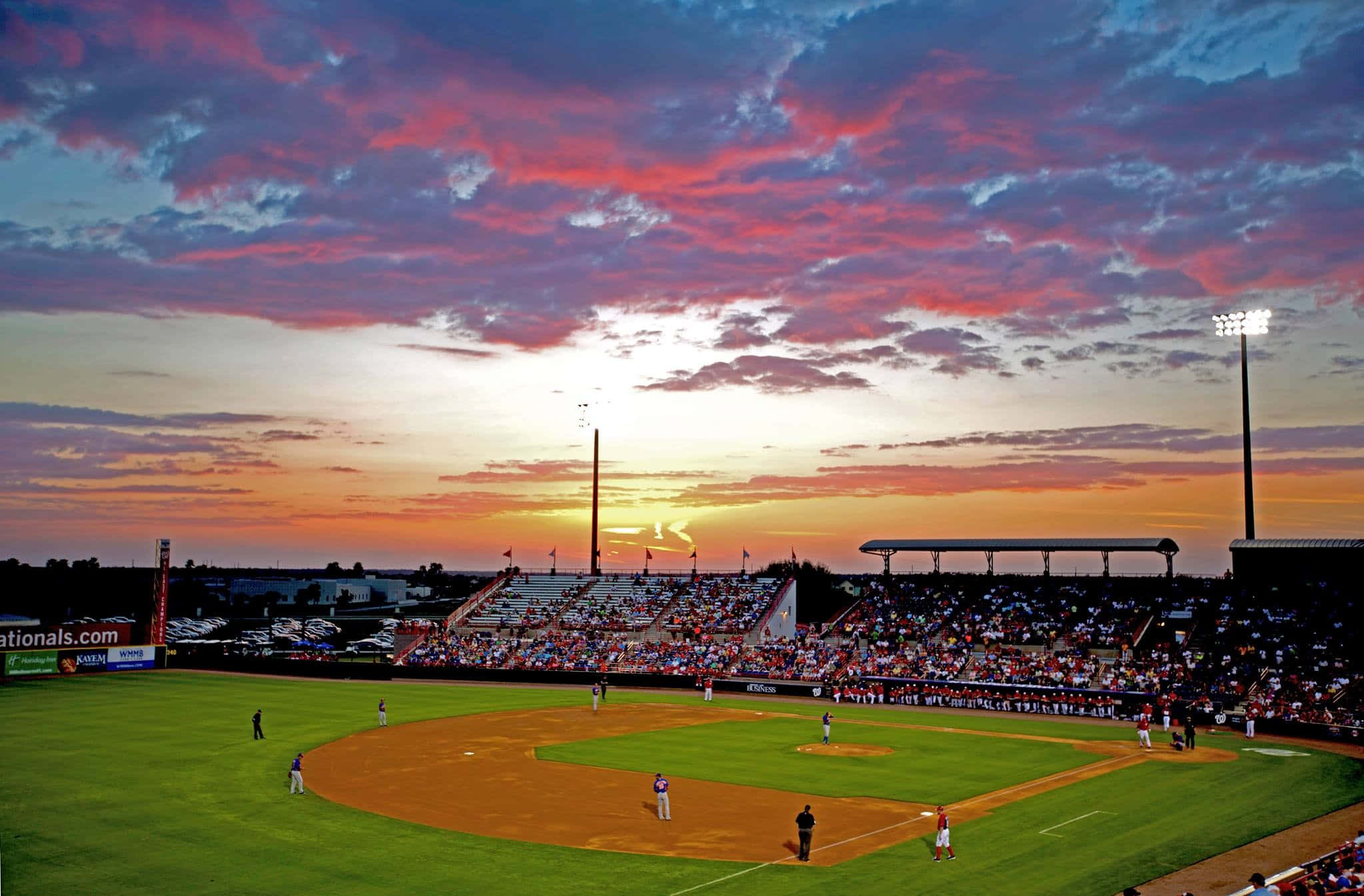 Baseball Field 桌布 壁紙 — 夕陽下的棒球場，場上有球員，看台上有球迷，天空呈現鮮豔的粉橙色。