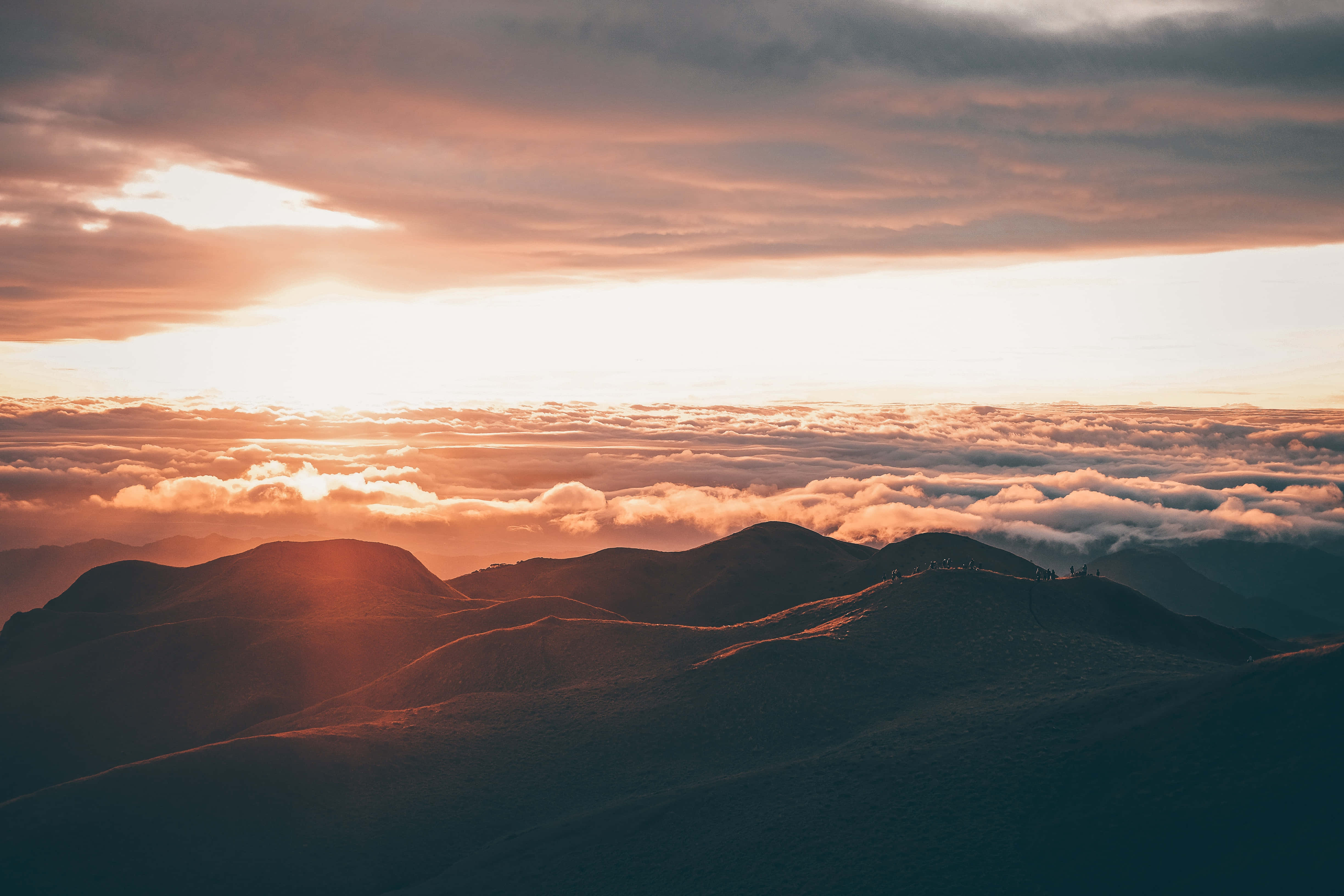 雲朵 桌布 壁紙 — 夕陽以溫暖的橙色光芒沐浴山頂，雲海之上山脊上徒步者的剪影。