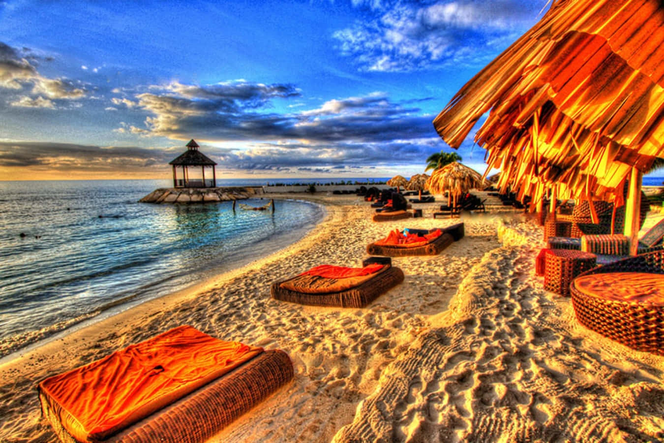 Jamaica 桌布 壁紙 — 夕陽以溫暖的橙色光芒沐浴著熱帶海灘，沙灘上排列著躺椅，旁邊是茅草屋頂的小屋和碼頭上的涼亭。