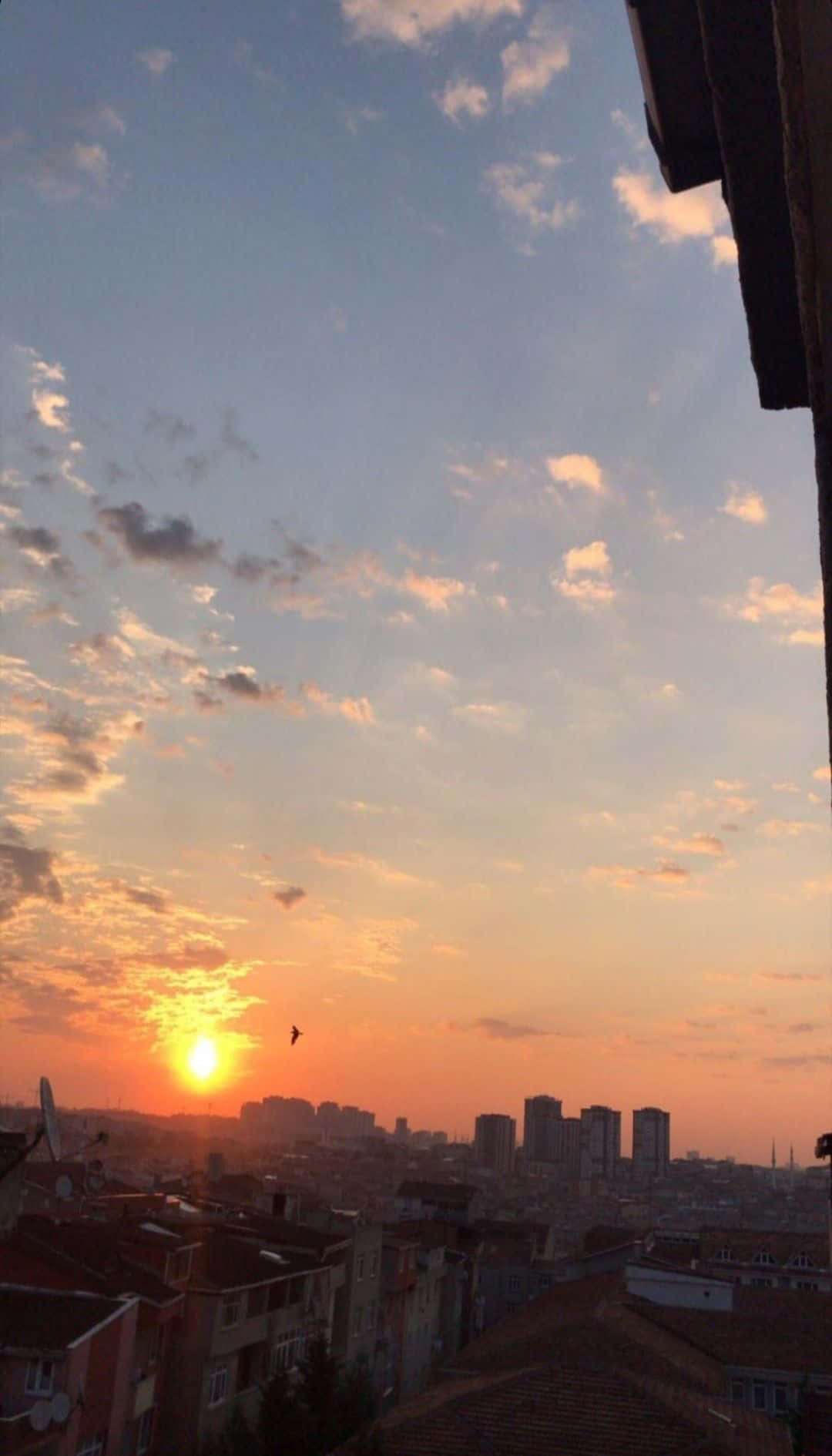 攝影 桌布 壁紙 — 夕陽以溫暖的橙色和粉紅色調描繪天空，映照著城市景觀中的屋頂和遠處的摩天大樓。