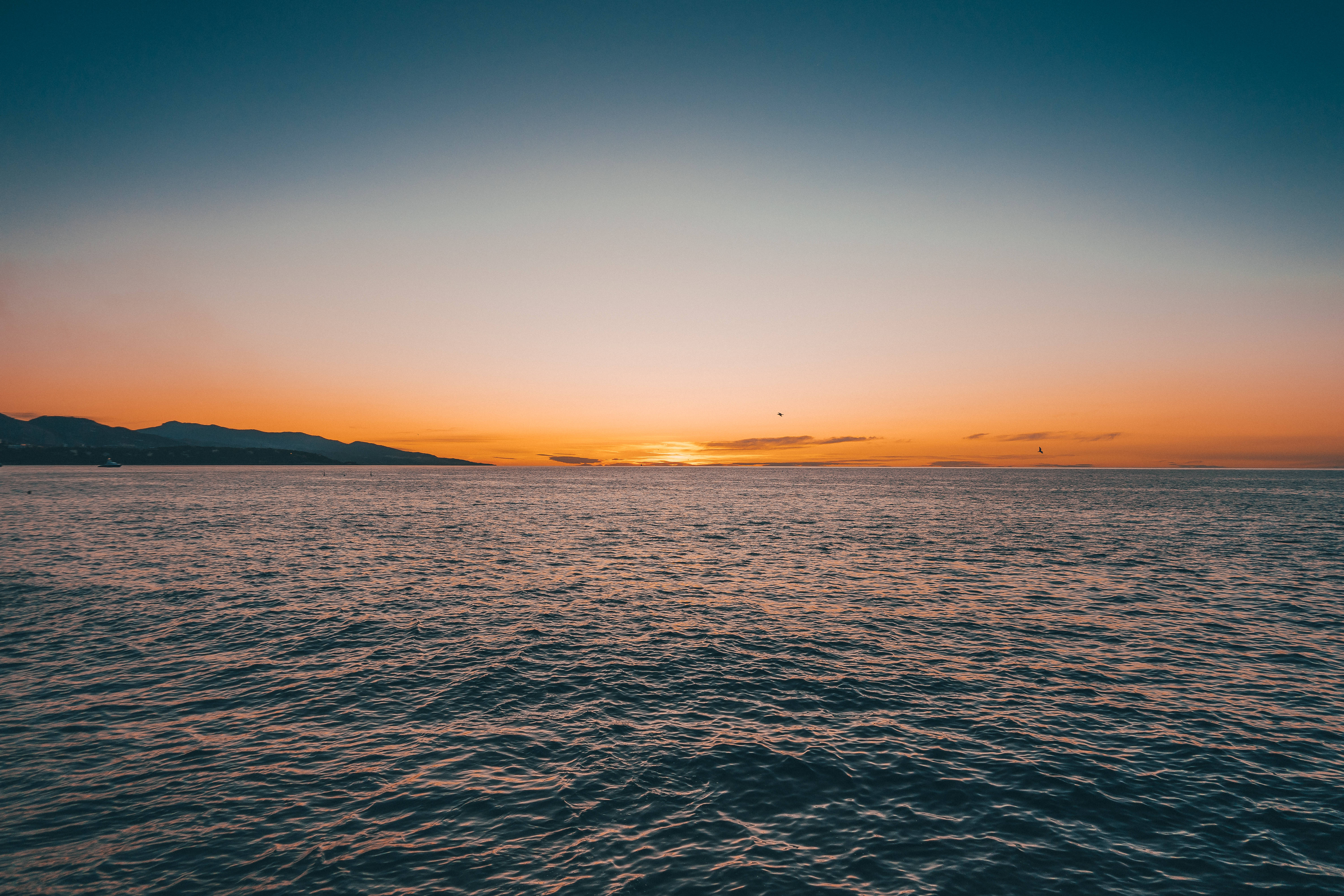 海洋 桌布 壁紙 — 夕陽以溫暖的橙色描繪地平線，沉落在遠山之後，映照在平靜的海面上。