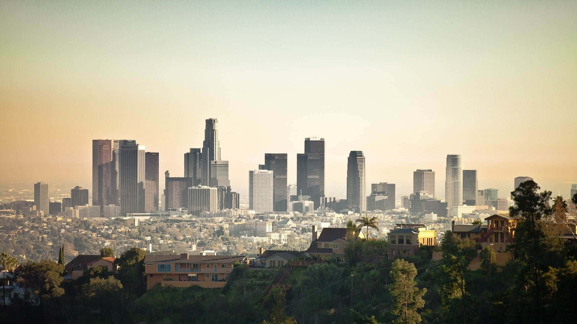 城市 桌布 壁紙 — 夕陽以溫暖的金色光芒沐浴洛杉磯的天際線，前景有棕櫚樹和住宅屋頂。