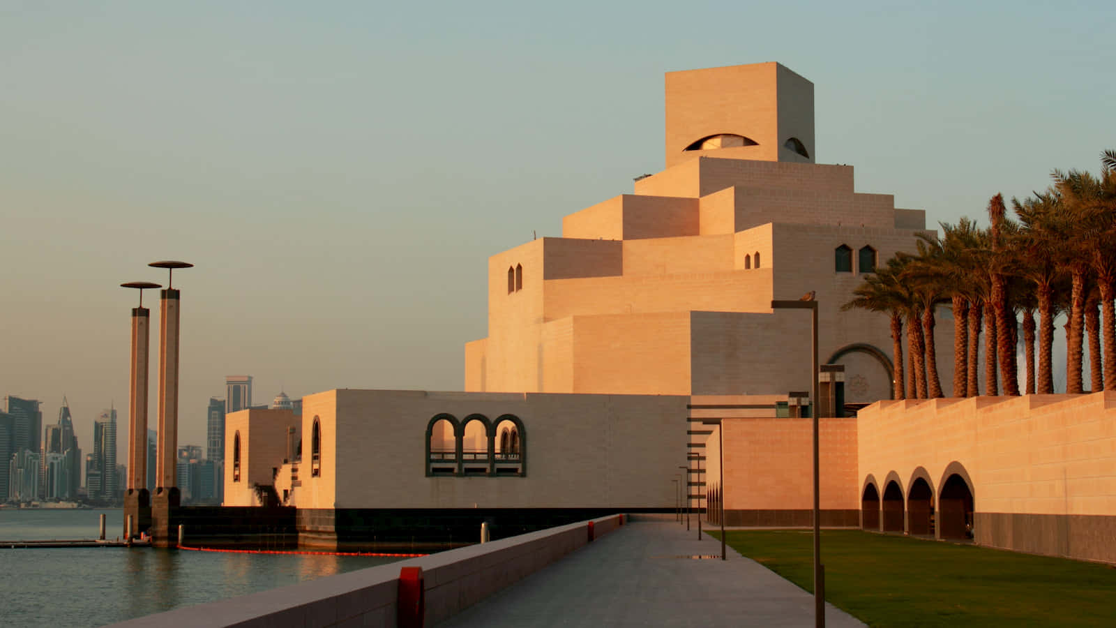 Museum Of Islamic Art 桌布 壁紙 — 夕陽以溫暖的金色光芒沐浴著伊斯蘭藝術博物館，棕櫚樹排列在其海濱步道兩旁，背景是城市天際線。