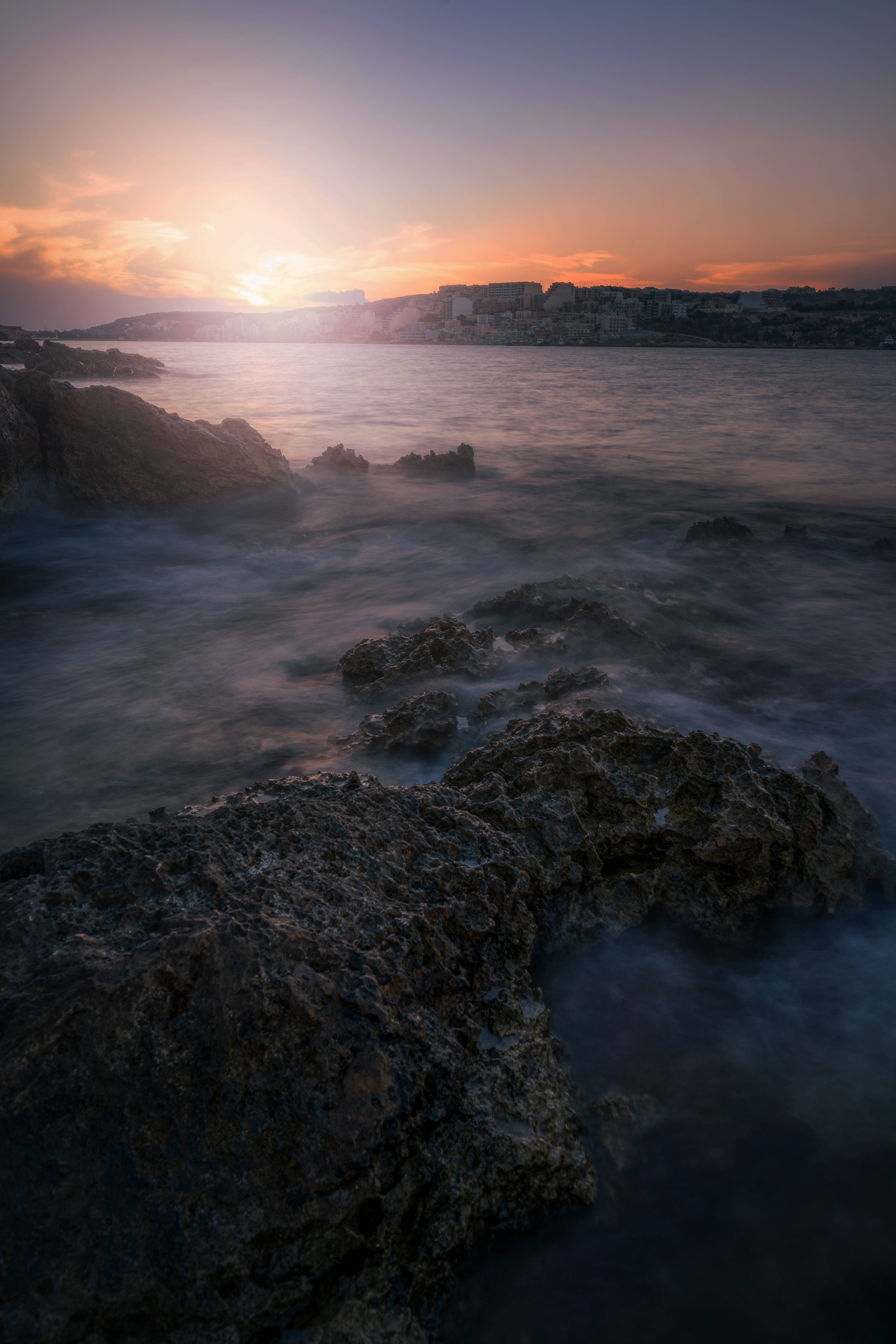 Malta 桌布 壁紙 — 夕陽以溫暖色調塗抹天空，柔和的海浪在崎嶇的海岸岩石周圍流動，遠方的小鎮在地平線上形成剪影。