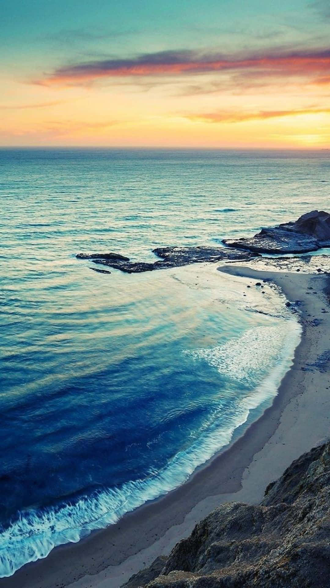 海灘 桌布 壁紙 — 夕陽以溫暖色調塗抹天空，海浪輕輕拍打著被崎嶇懸崖環繞的隱蔽沙灘。