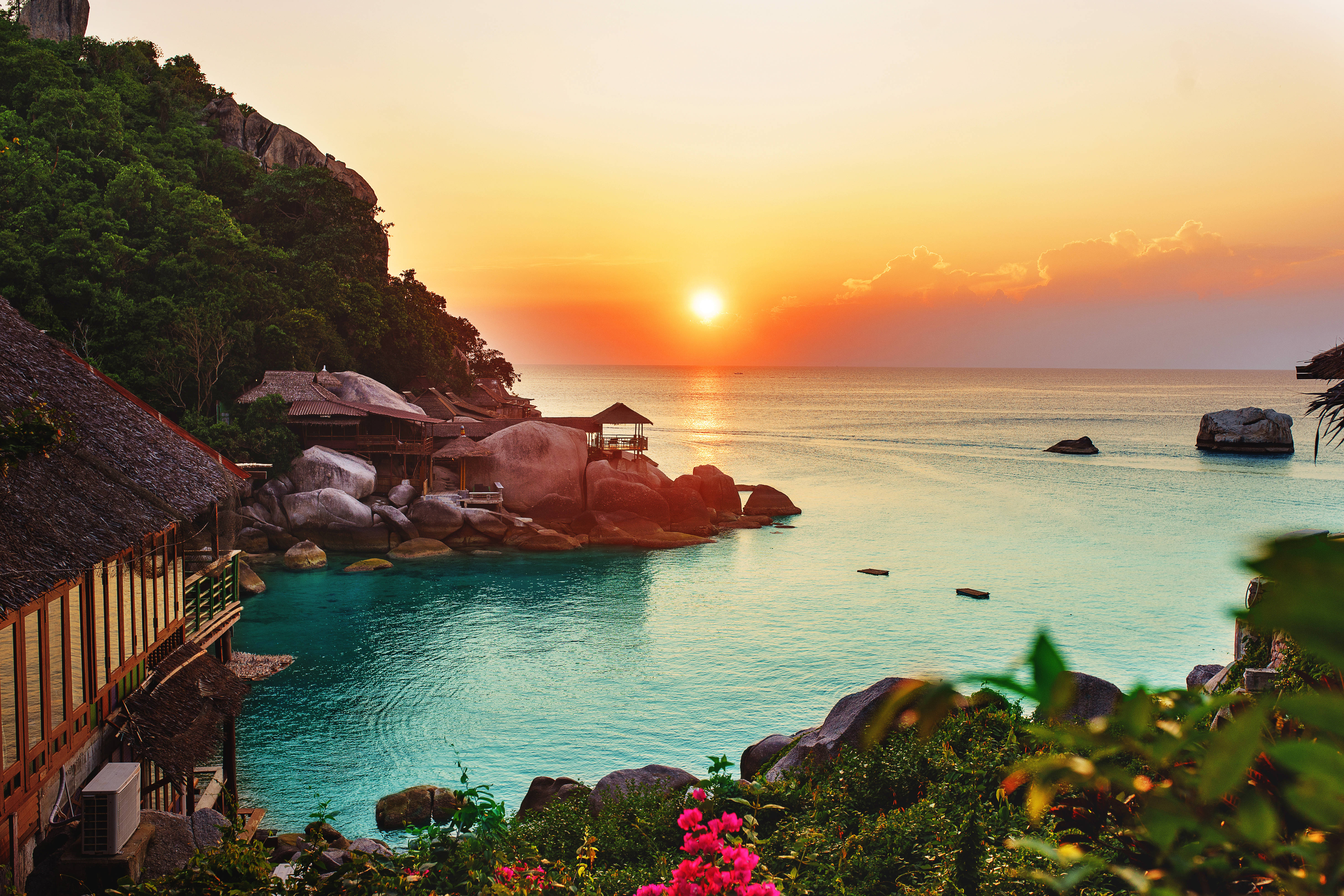 海洋 桌布 壁紙 — 夕陽以溫暖色調沐浴熱帶海灣，木屋坐落在茂密綠植與碧綠海水之間。