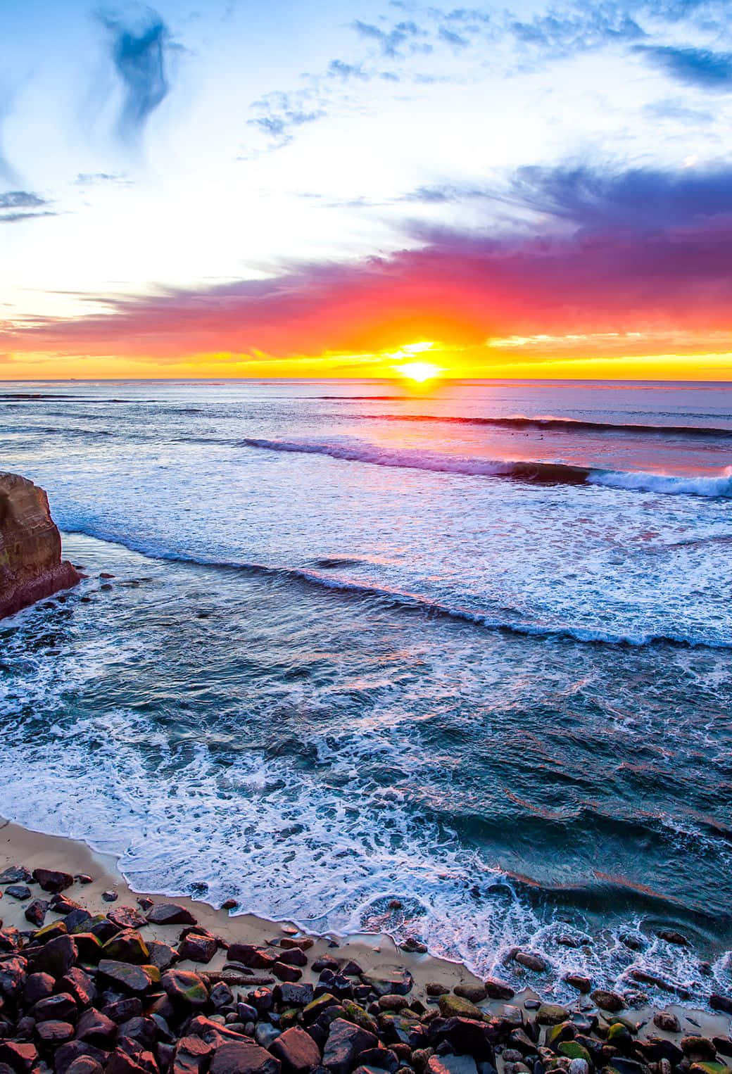 San Diego iPhone 桌布 壁紙 — 夕陽以火熱的橙色和粉紅色調為海洋塗上鮮豔色彩，海浪拍打岩石海岸。