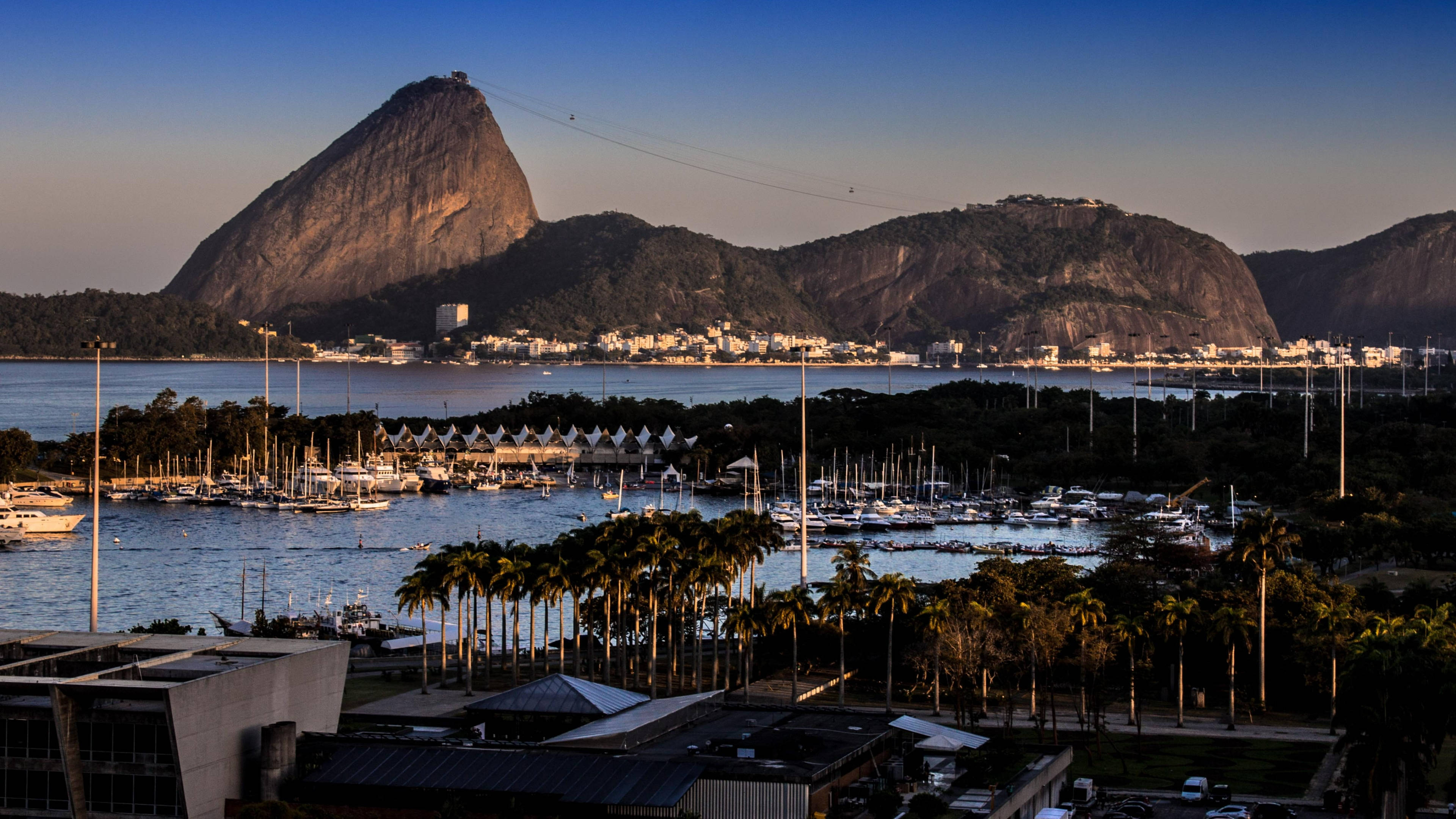 rio 桌布 壁紙 — 夕陽以金色光芒沐浴著糖麵包山和繁忙的碼頭，棕櫚樹和停泊的船隻點綴其間。