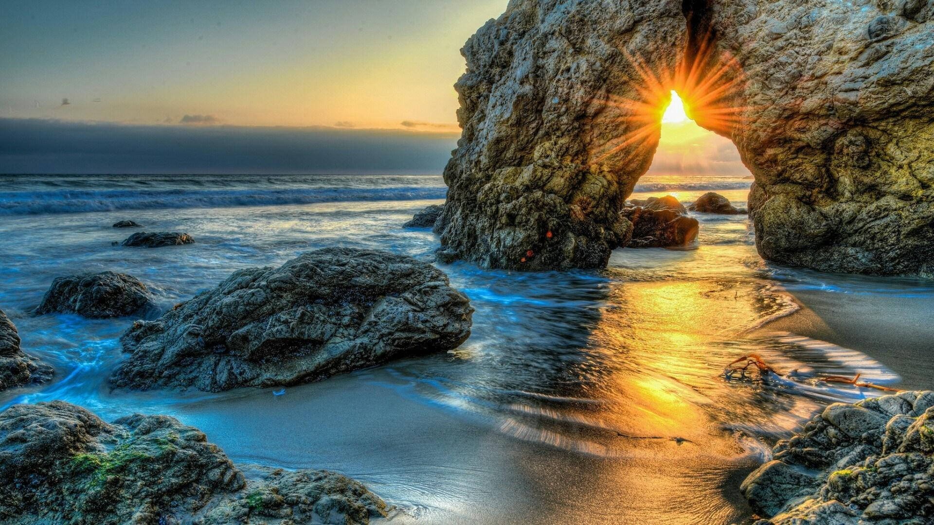 rocky 桌布 壁紙 — 夕陽光芒穿過天然岩石拱門，海浪拍打岩石海灘，泛起金色倒影。