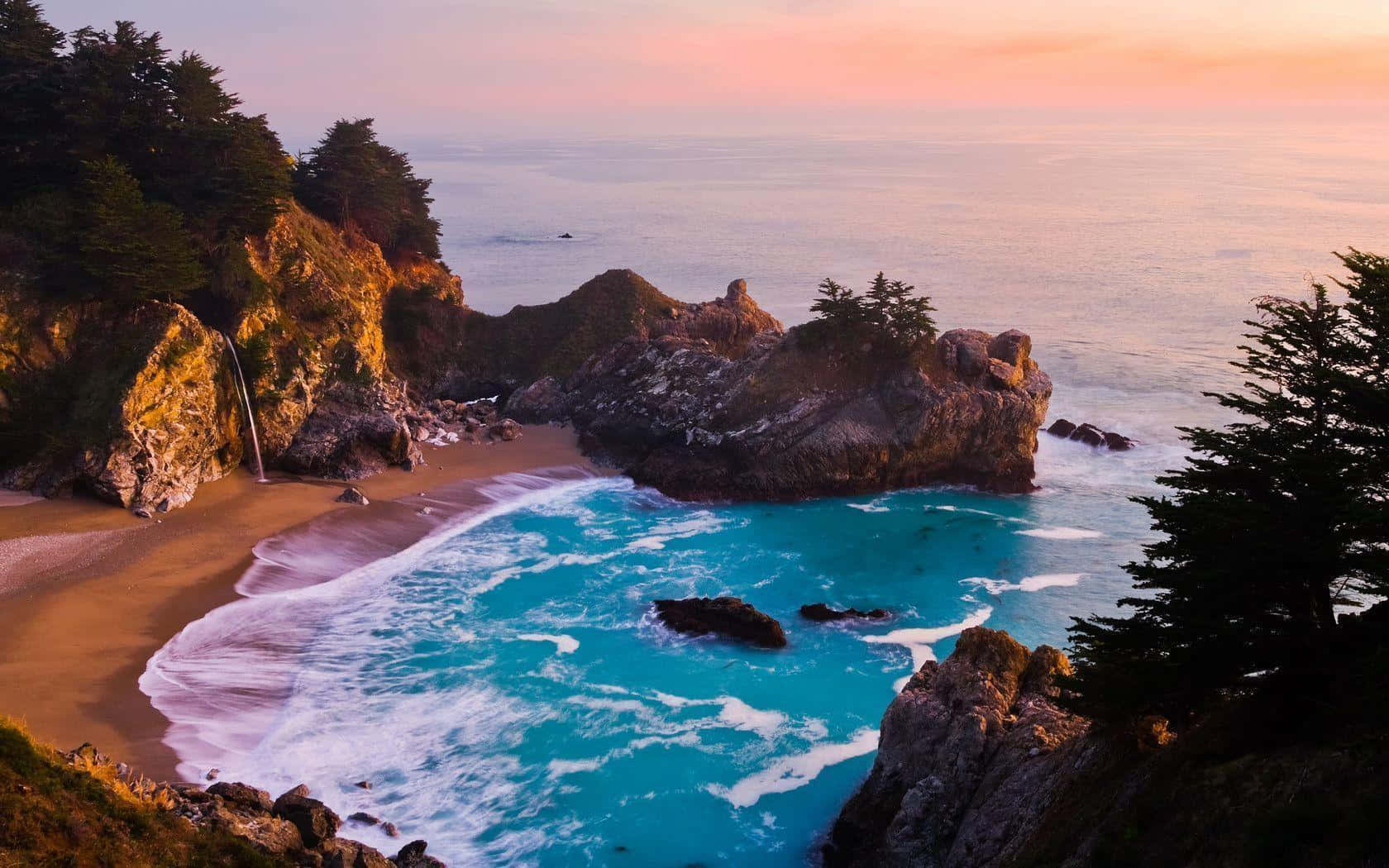 Big Sur 桌布 壁紙 — 夕陽在一個僻靜的海灣上空繪出鮮豔的天空，碧綠的海水拍打著金色的沙灘和崎嶇的懸崖。
