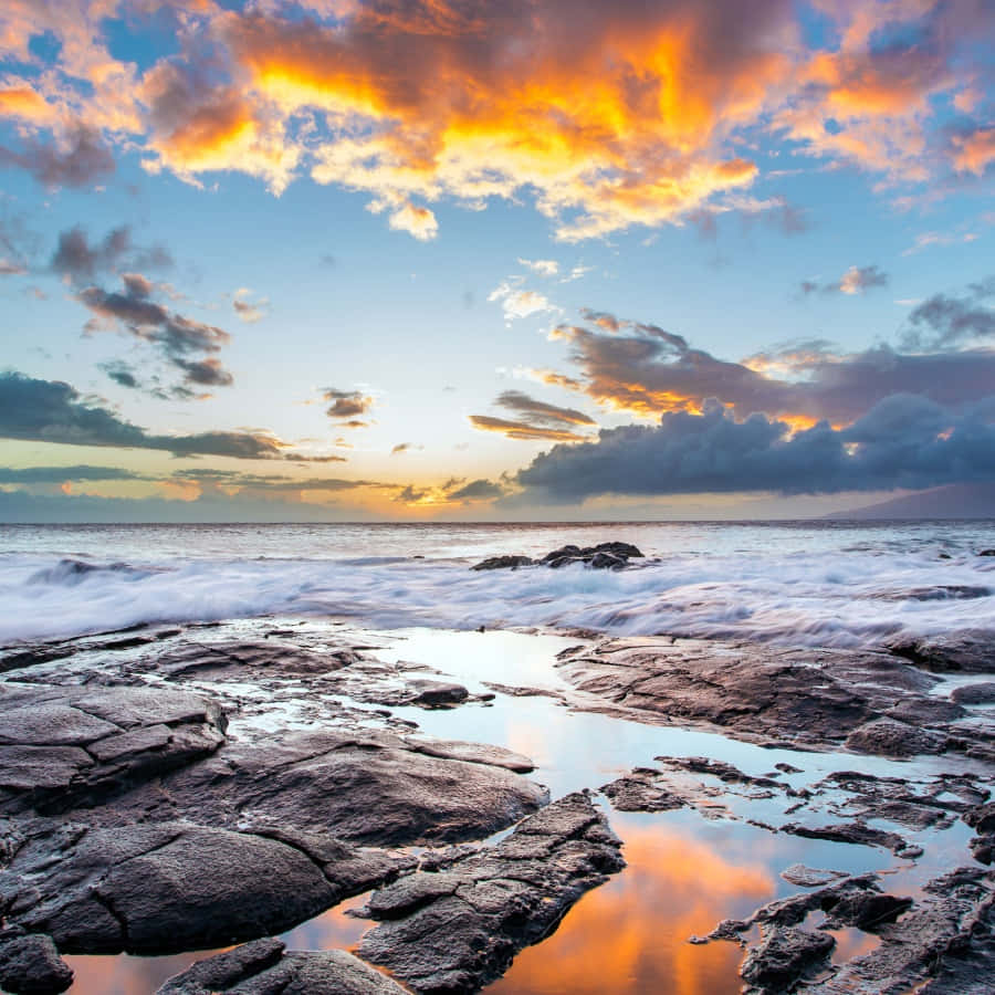 美學 iPad 壁紙 桌布 壁紙 — 夕陽在岩石海岸線上繪出火熱的橙色雲彩，海浪拍打並反射天空鮮豔的色彩。