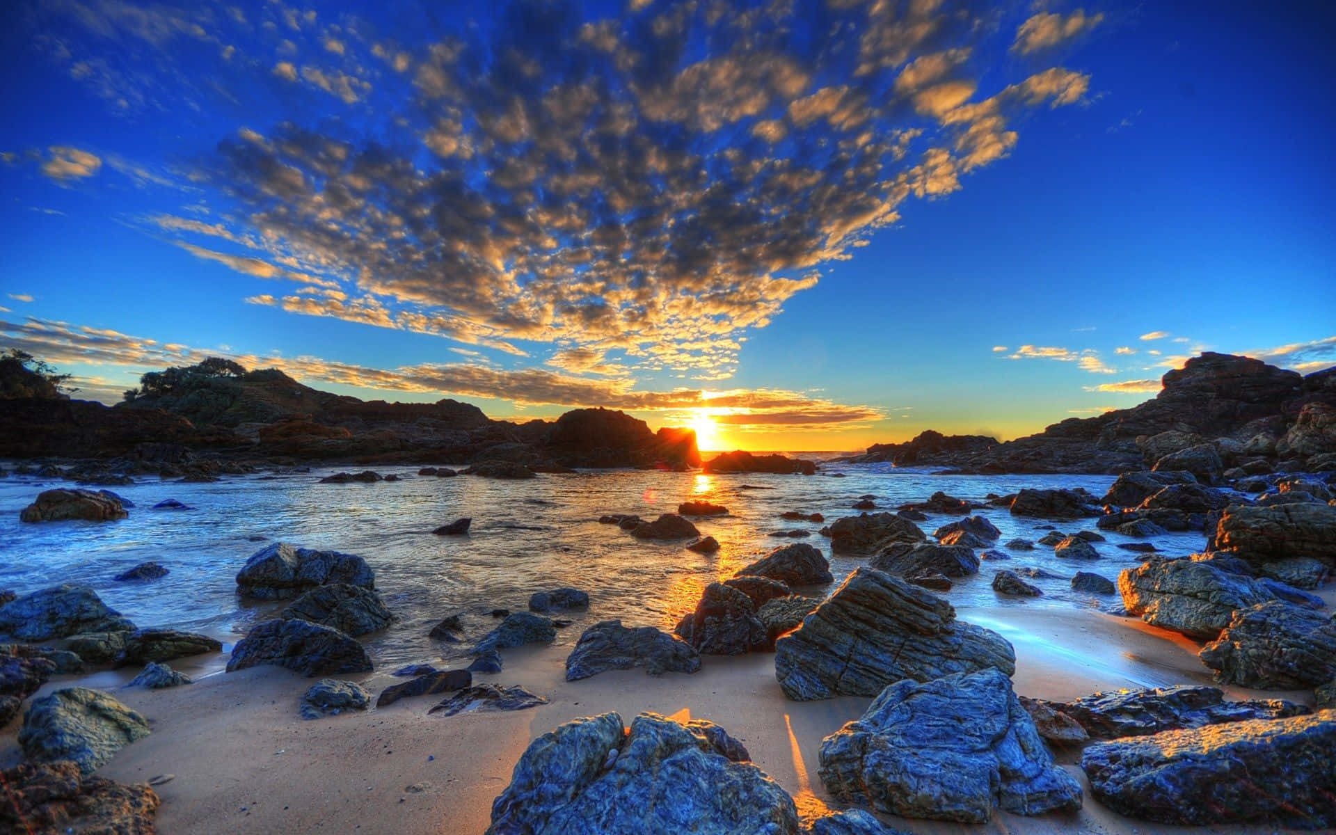 澳洲 桌布 壁紙 — 夕陽在岩石海灘上繪出金色雲彩，海浪輕輕拍打石頭和沙灘。