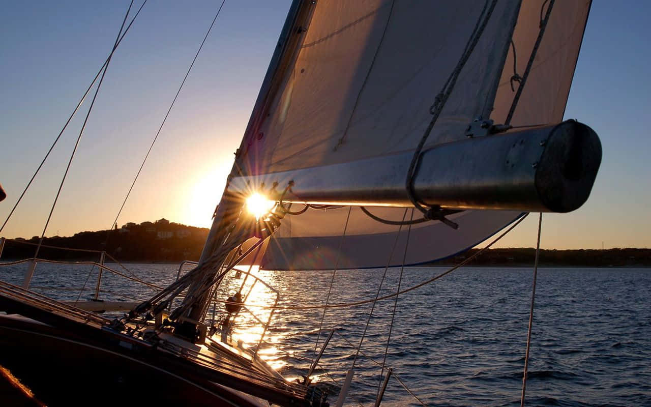 Sailboat 桌布 壁紙 — 夕陽在帆船的桅杆後發光，金色的光芒反射在漣漪的水面和遠處的海岸線上。