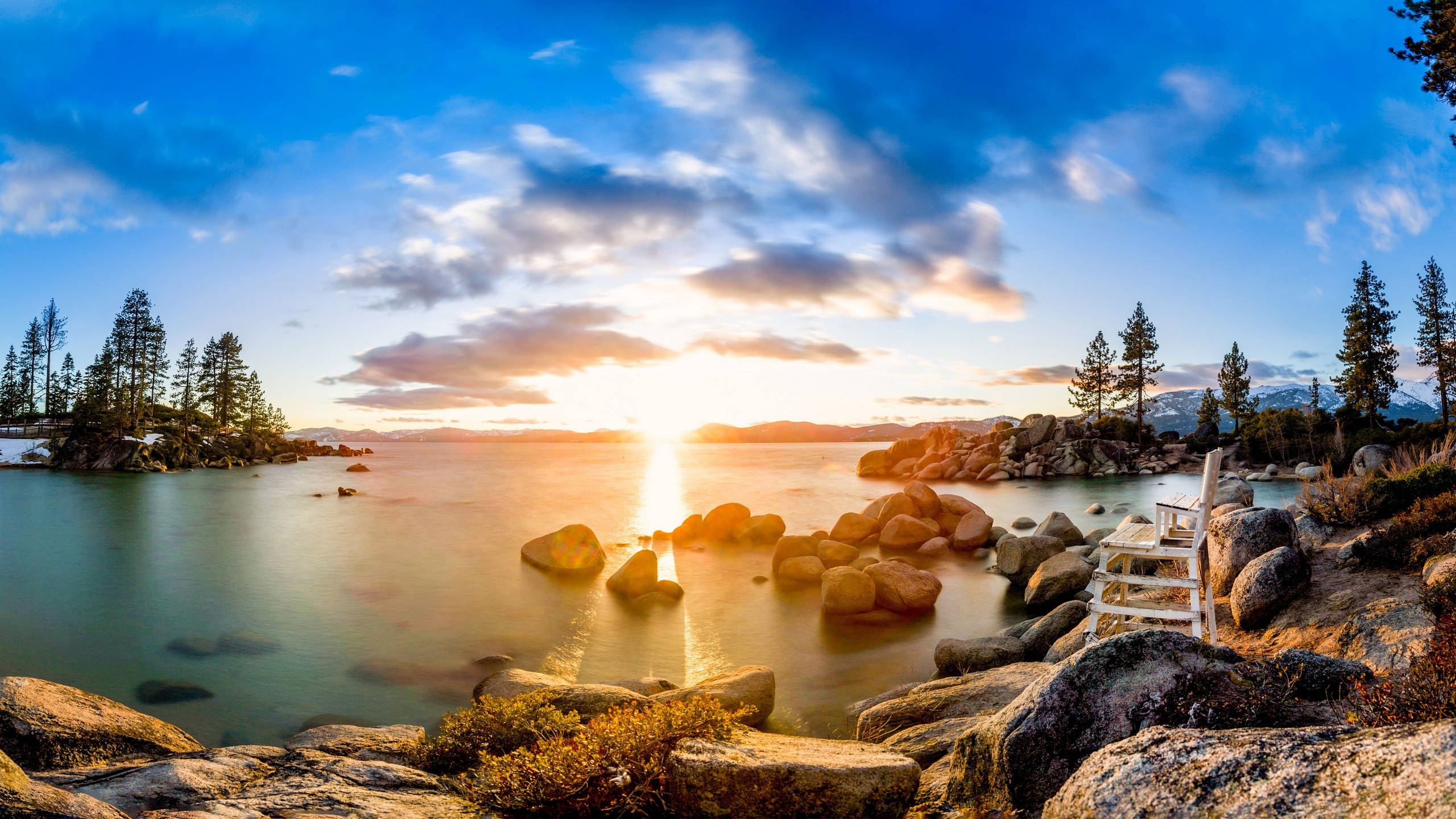 2560x1440 nature 桌布 壁紙 — 夕陽在松樹和岩石海岸環繞的寧靜湖面上閃耀，一張孤獨的椅子邀請靜思。