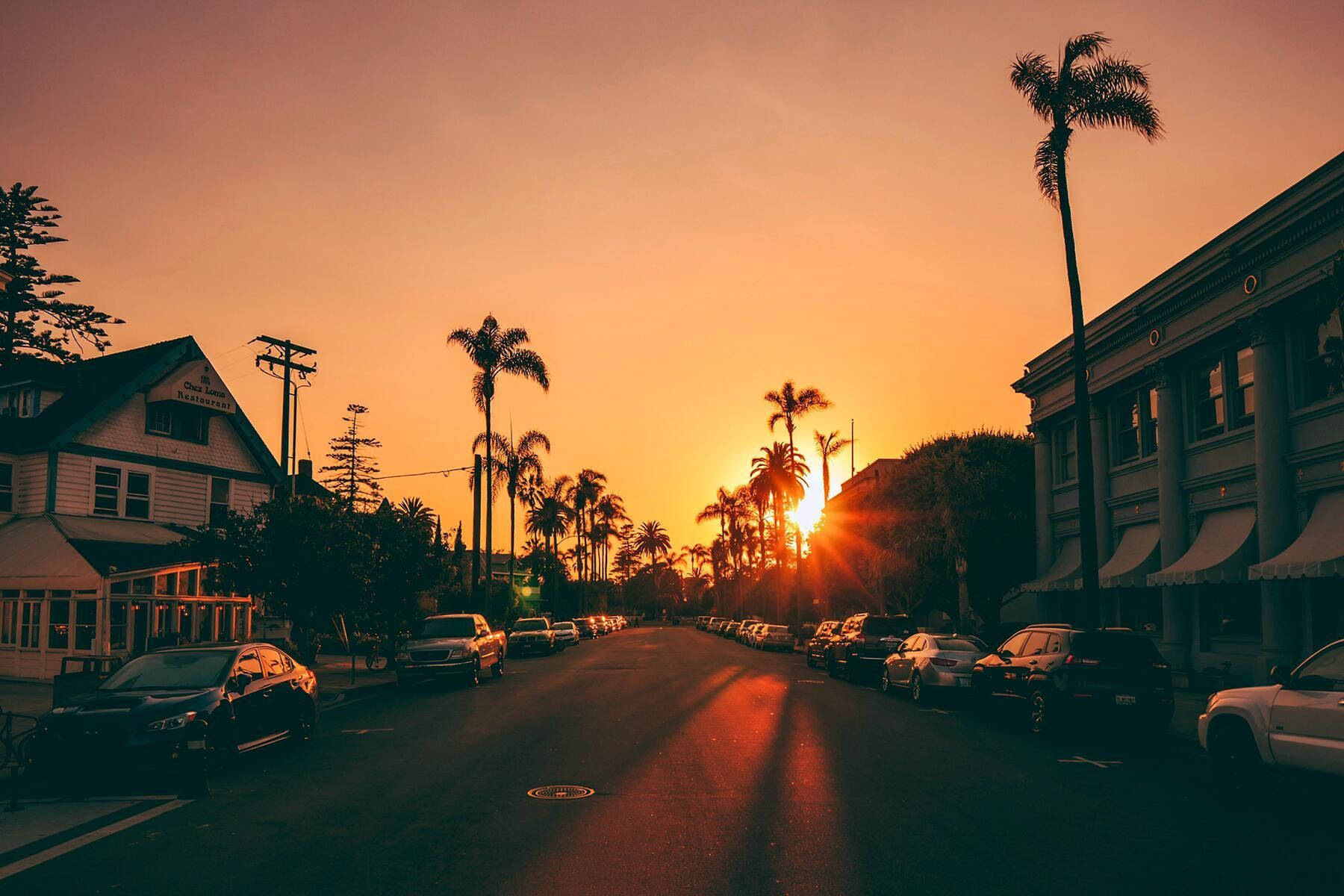Road 桌布 壁紙 — 夕陽在棕櫚樹成蔭的街道上投射出溫暖的橙色光芒，街道上停著汽車和復古建築。