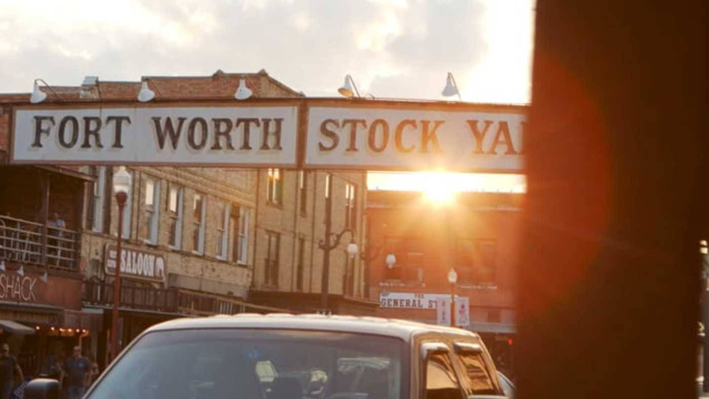 Worth 桌布 壁紙 — 夕陽在歷史悠久的Fort Worth Stockyards標誌後方發光，一輛汽車駛過復古建築。